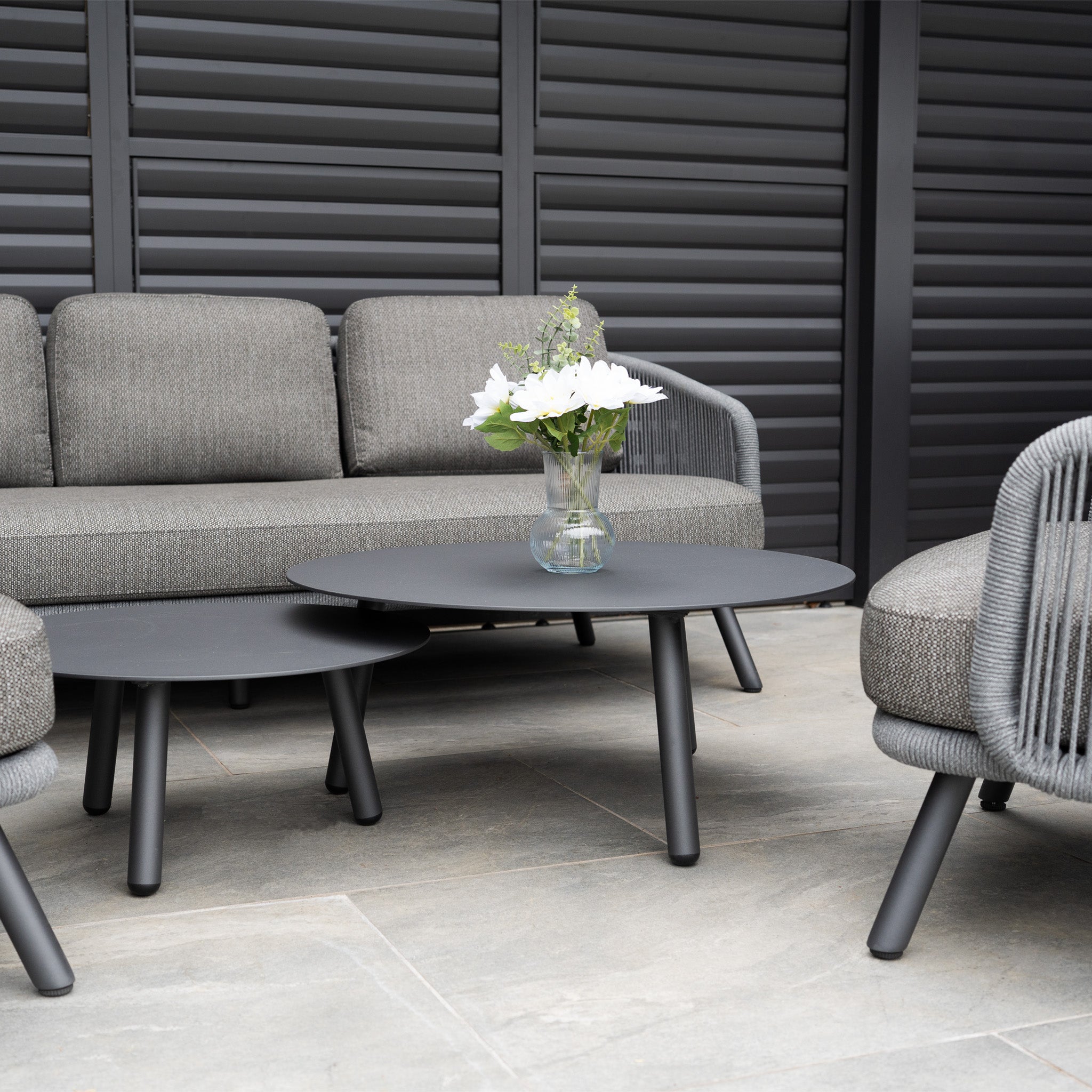 The Dekota 3 Seat Rope Sofa Set with Coffee Tables and Footstool in Charcoal offers outdoor luxury; the round table displays a vase of white flowers on a stone patio.