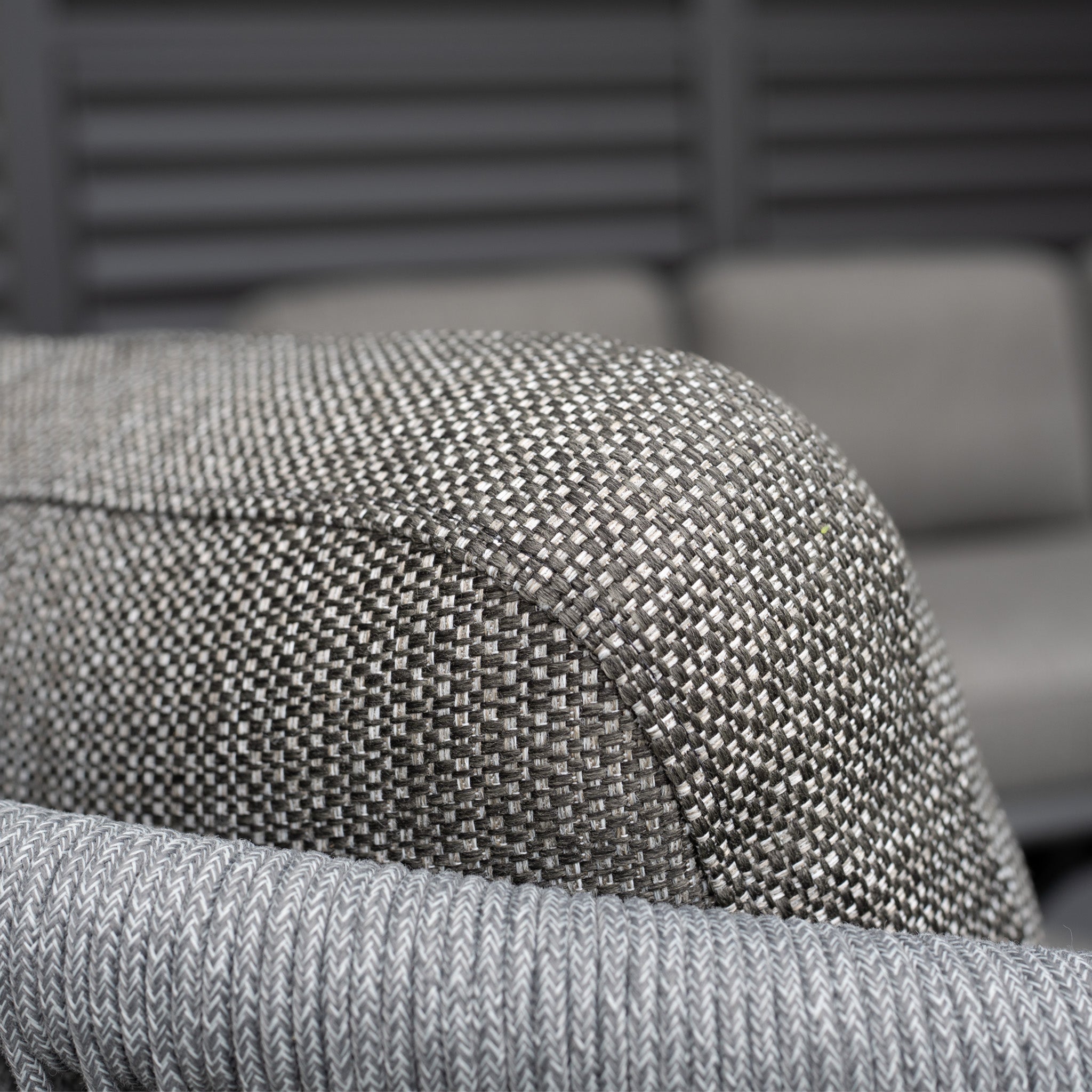 Close-up of the Dekota 3 Seat Rope Sofa Set with Coffee Tables and Footstool in Charcoal featuring a textured gray olefin fabric armrest, with a blurred matching sofa set in the background—highlighting outdoor luxury.