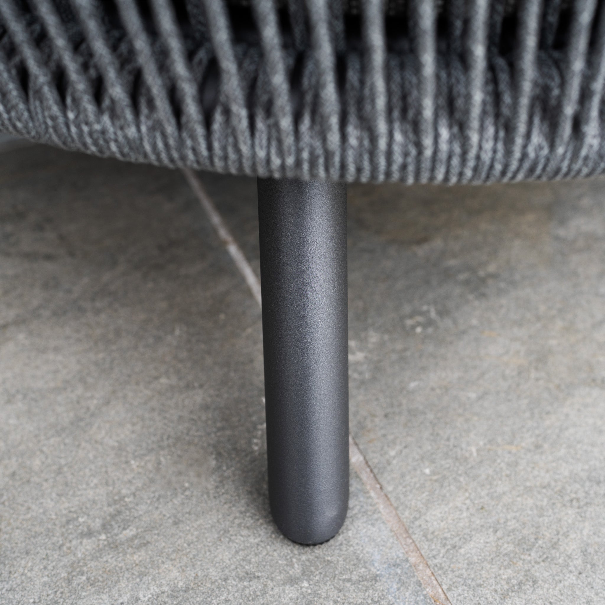Close-up of a black metal chair leg on gray tiles, topped with woven gray olefin fabric—perfect for adding outdoor luxury to your Dekota 3 Seat Rope Sofa Set with Coffee Tables and Footstool in Charcoal.