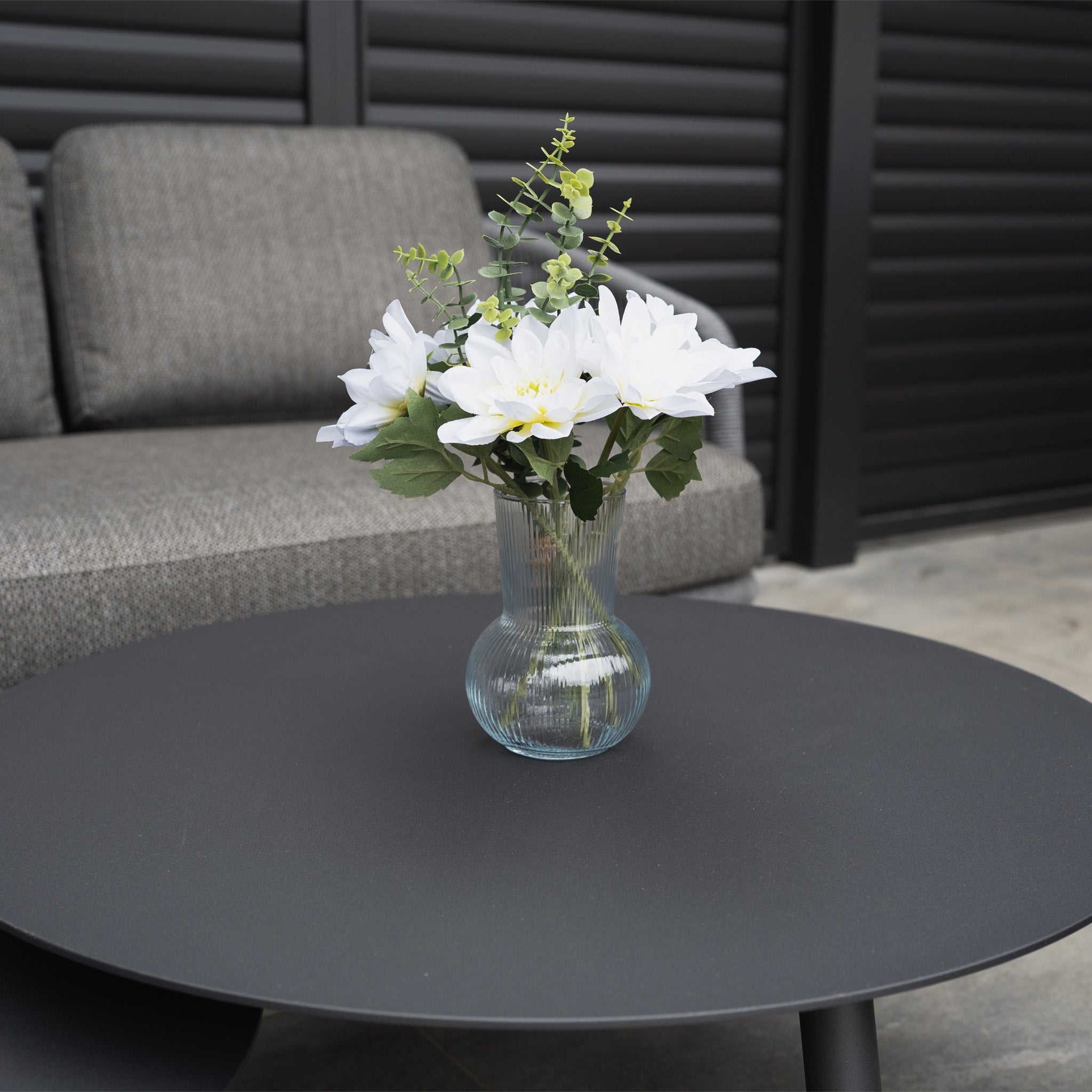 A glass vase with white flowers and greenery sits on a round black table before the Dekota 3 Seat Rope Sofa Set with Coffee Tables and Footstool in Charcoal, creating a stylish outdoor setting.