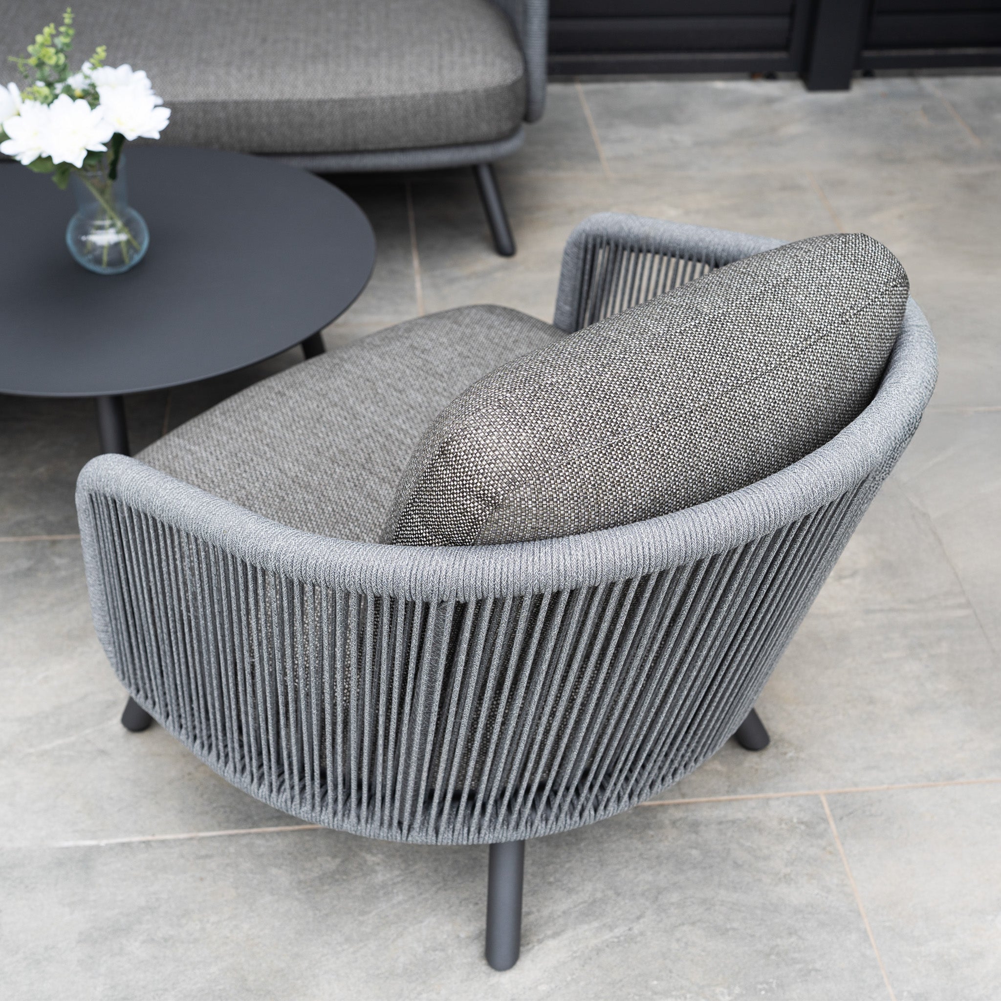 A modern gray lounge chair with rope detailing and olefin fabric sits by a round black table with a white vase, creating outdoor luxury that perfectly complements the Dekota 3 Seat Rope Sofa Set with Coffee Tables and Footstool in Charcoal.