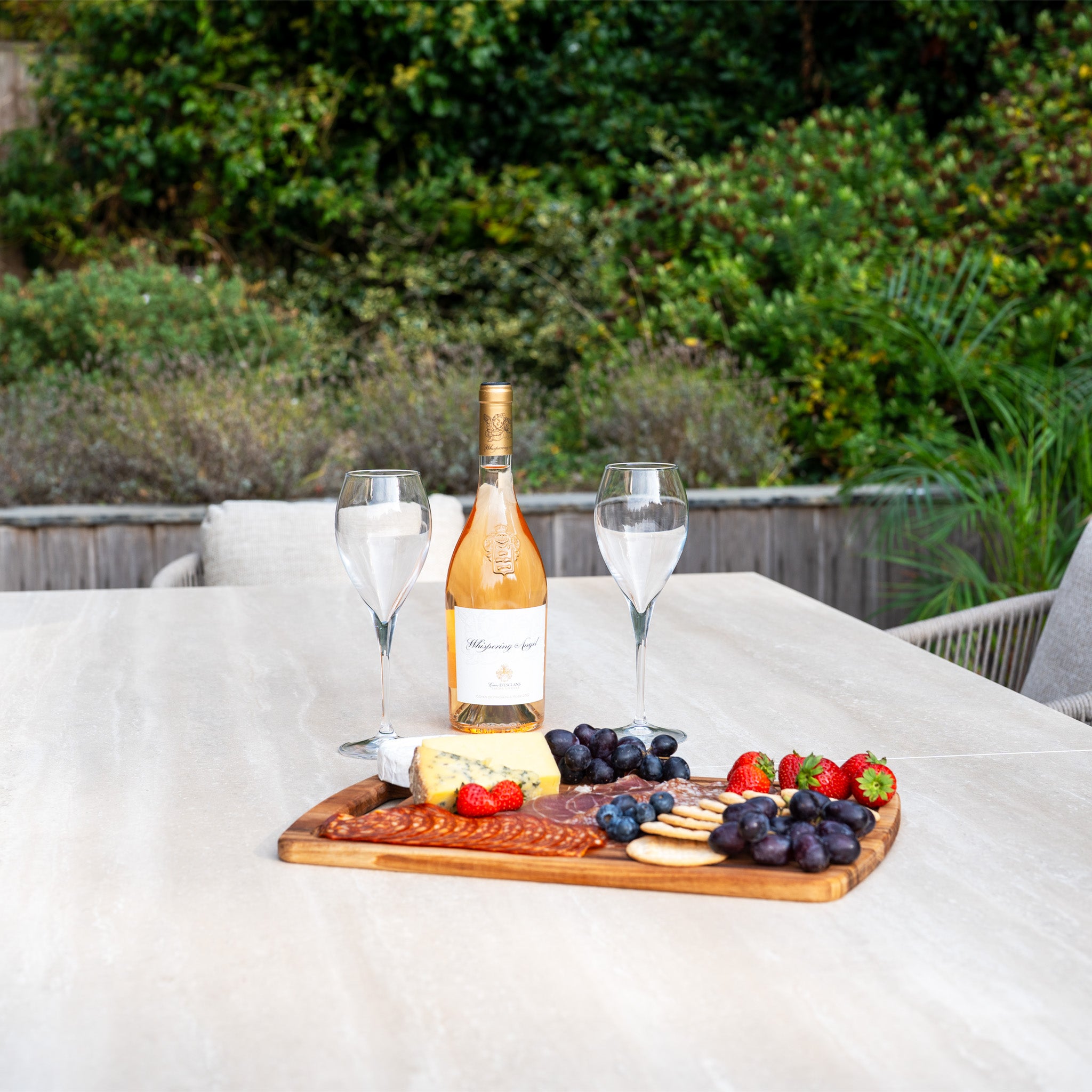 A bottle of wine, two glasses, and a cheese and fruit platter rest on the Dekota 6 Seat Rope Dining Set with Albatross Extending Table in Fawn, surrounded by lush greenery.