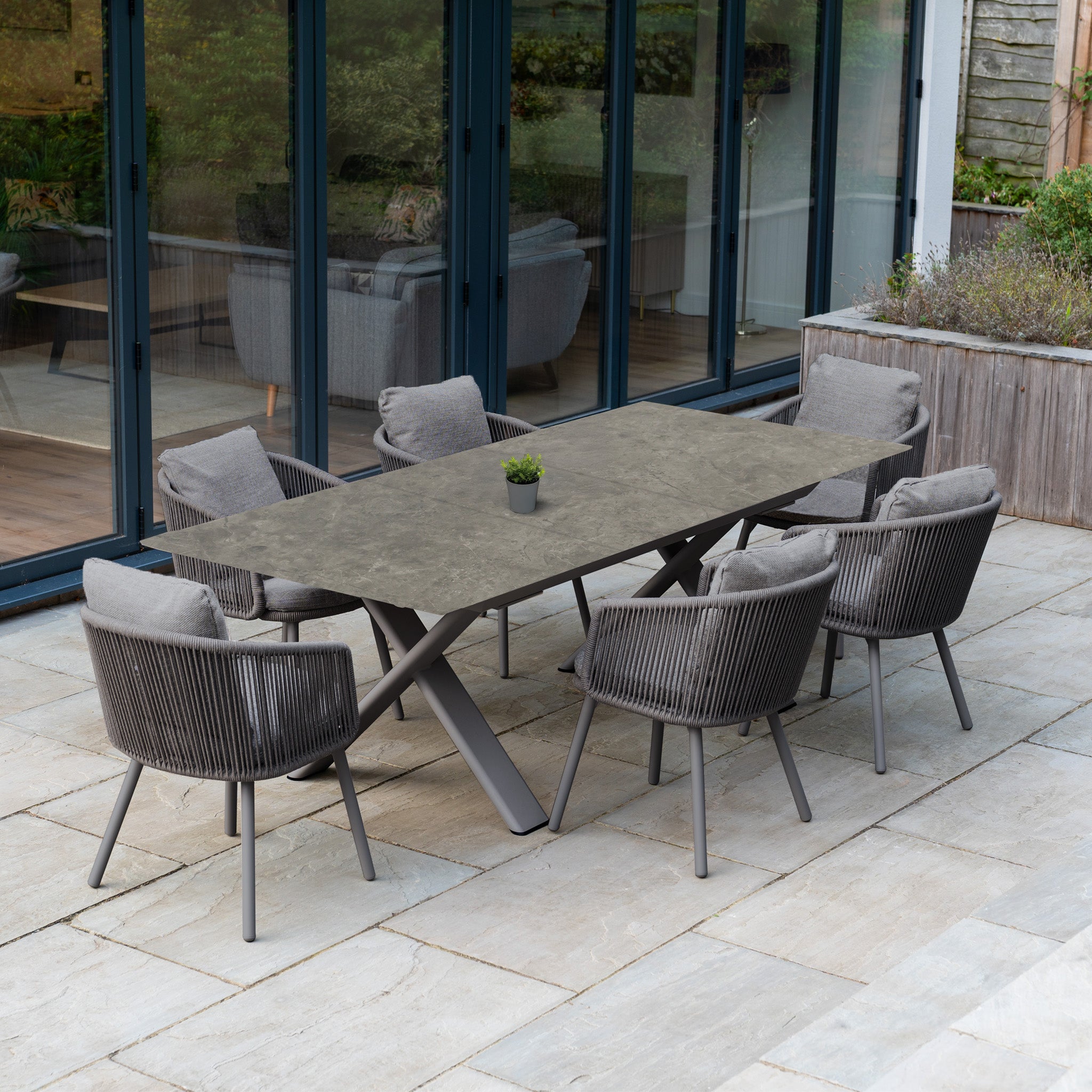 The Dekota 6 Seat Rope Dining Set with Albatross Extending Table in Charcoal features six cushioned chairs on a stylish patio, complete with a small plant centerpiece.