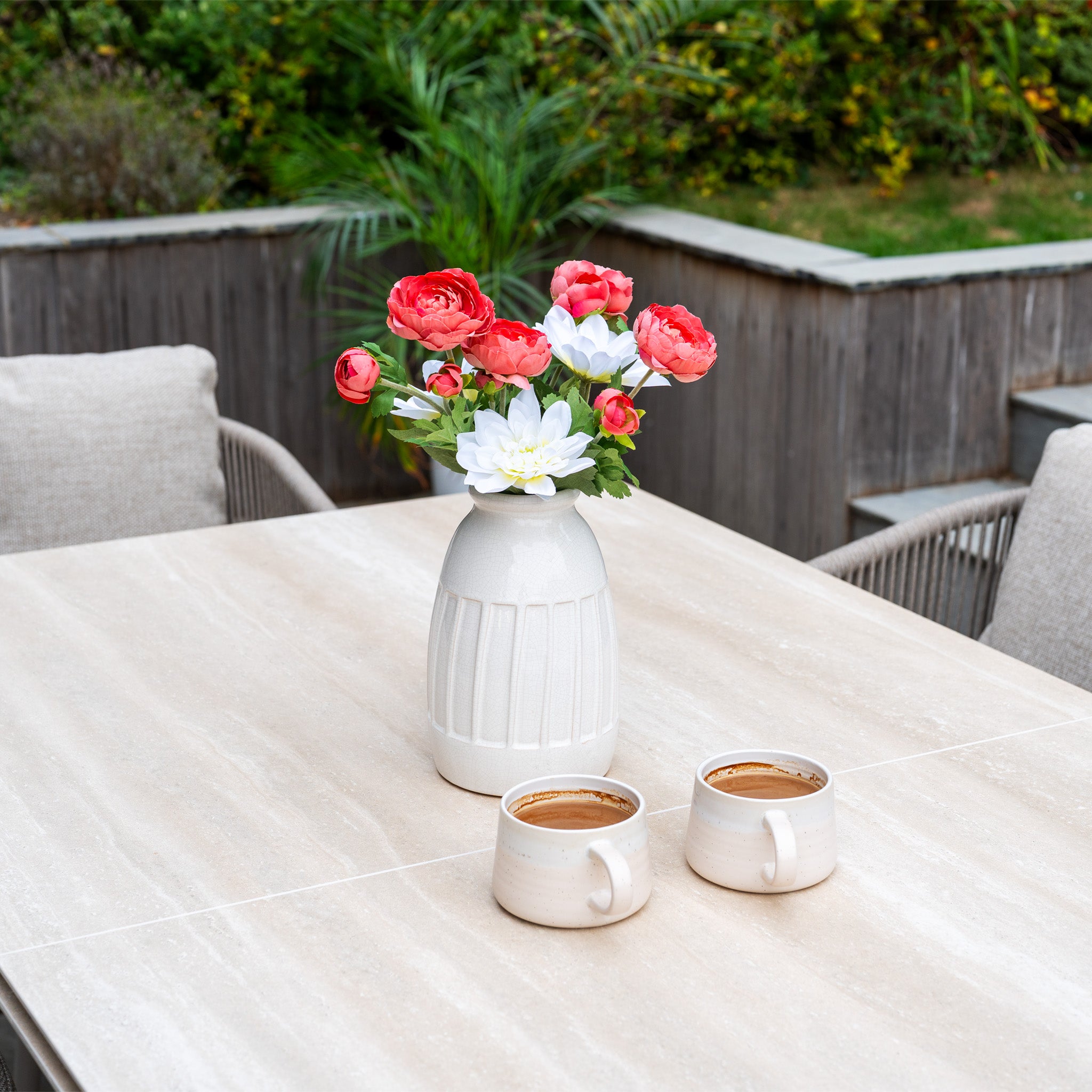 A white vase with red and white flowers and two coffee mugs sit on the Dekota 6 Seat Rope Dining Set with Albatross Extending Table in Fawn, surrounded by greenery—ideal for relaxing mornings or gatherings.