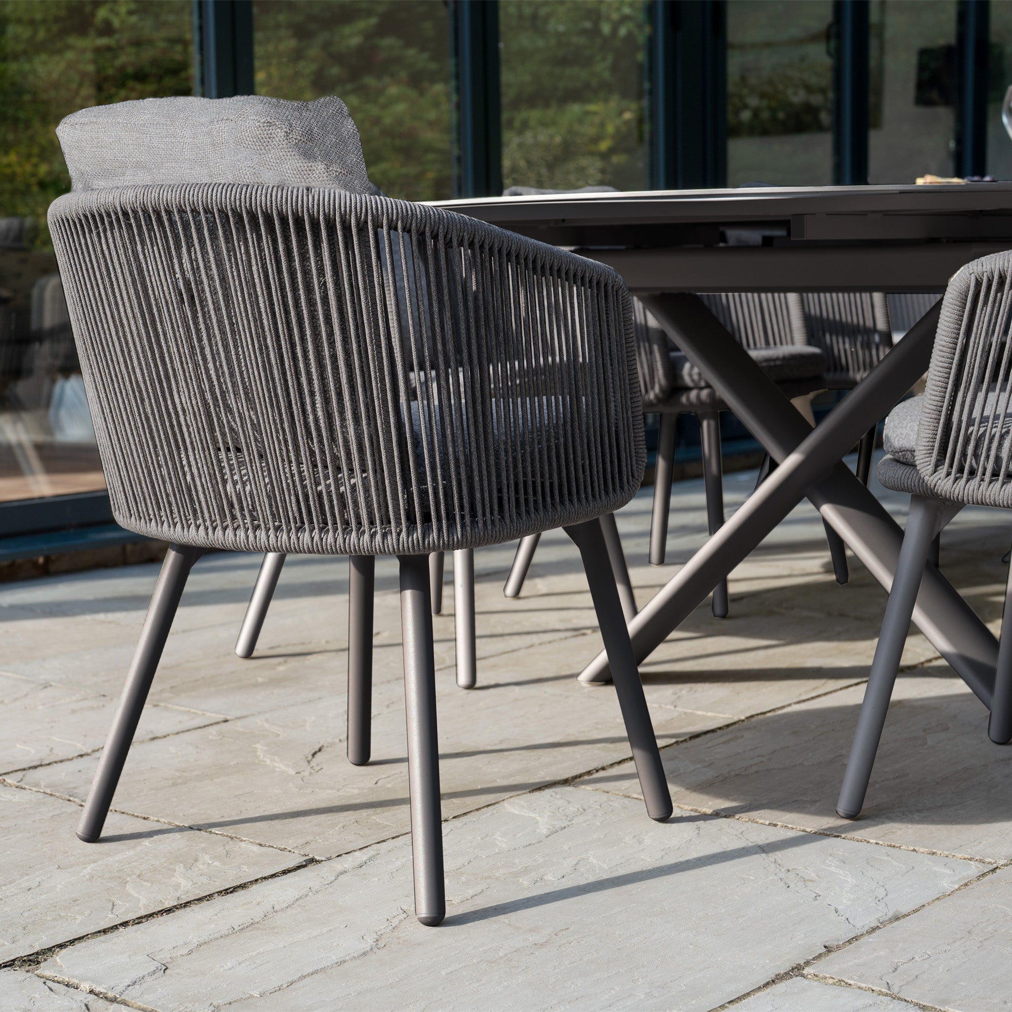 The Dekota 8 Seat Oval Rope Dining Set with Albatross Extending Table in Charcoal comfortably seats eight, ideal for gatherings on a stone patio surrounded by lush greenery and windows.
