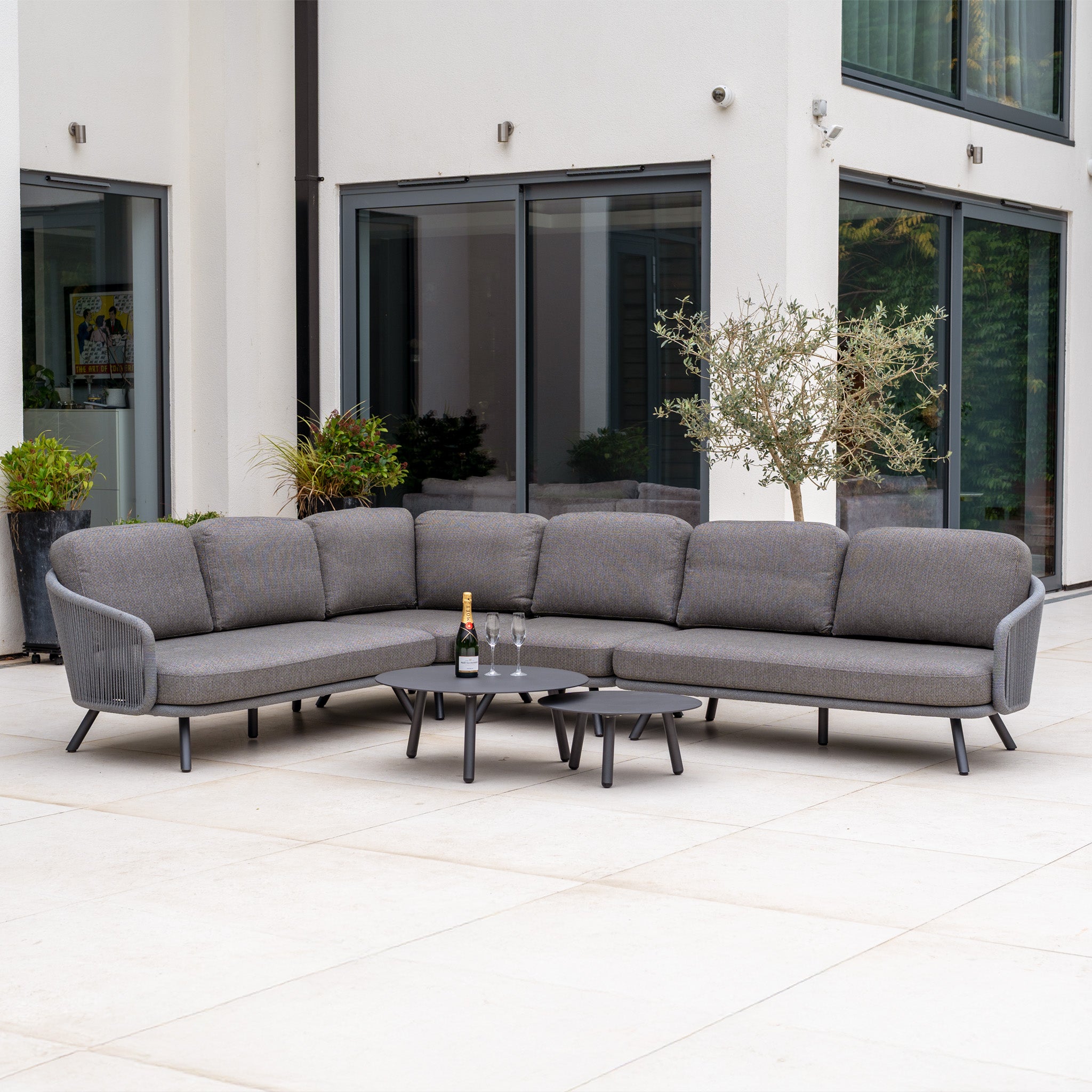 Dekota Large Corner Sofa Set in Charcoal with coffee tables, champagne, and glasses on a spacious patio by a modern building—stylish outdoor furniture for elegant entertaining.