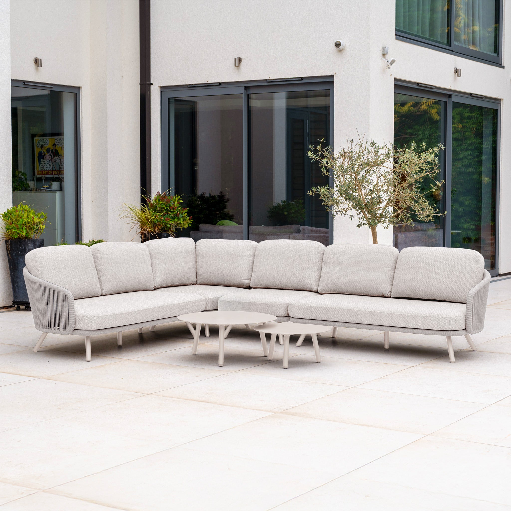 Dekota Large Corner Sofa Set in Fawn with hydrophobic rope weave and coffee table on a patio with plants, placed before large windows.