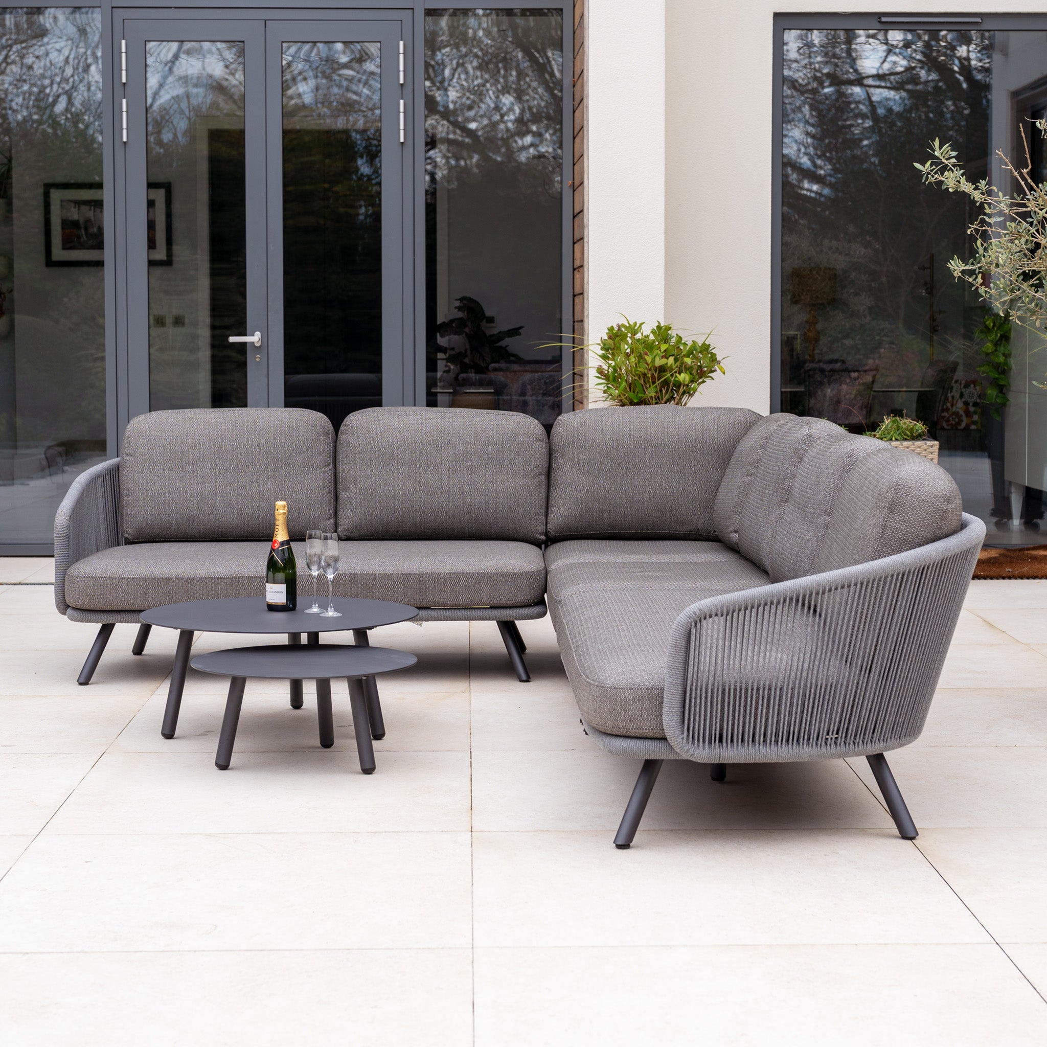 Dekota Large Corner Sofa Set in Charcoal with a round coffee table, champagne bottle, and glasses on a patio—ideal for relaxing with stylish outdoor furniture.