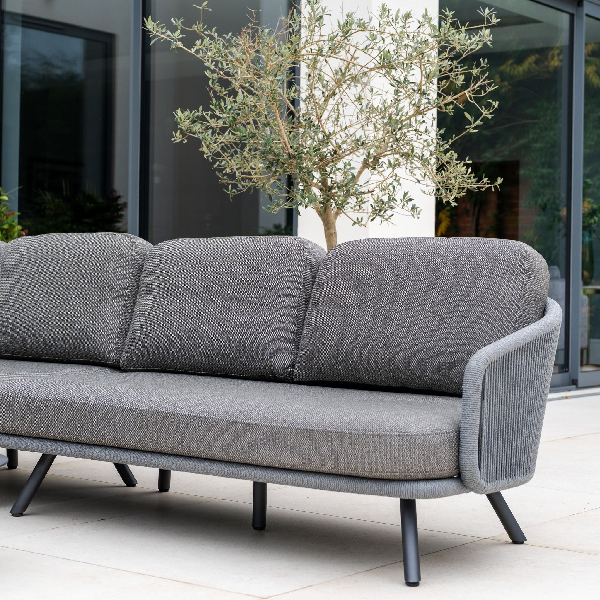 The Dekota Large Corner Sofa Set in Charcoal is placed on a patio near a glass window, featuring three cushions and a small potted tree, creating a stylish outdoor setting.