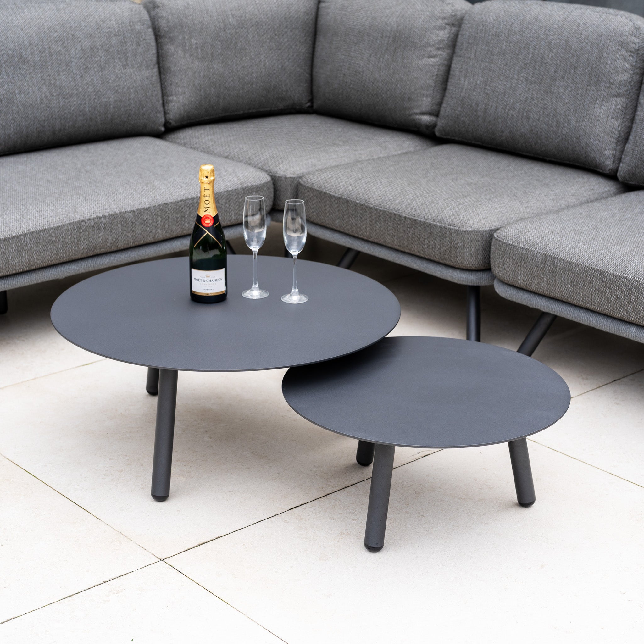 Two round black coffee tables with champagne and two glasses are paired with the Dekota Large Corner Sofa Set in Charcoal for a stylish outdoor furniture arrangement.