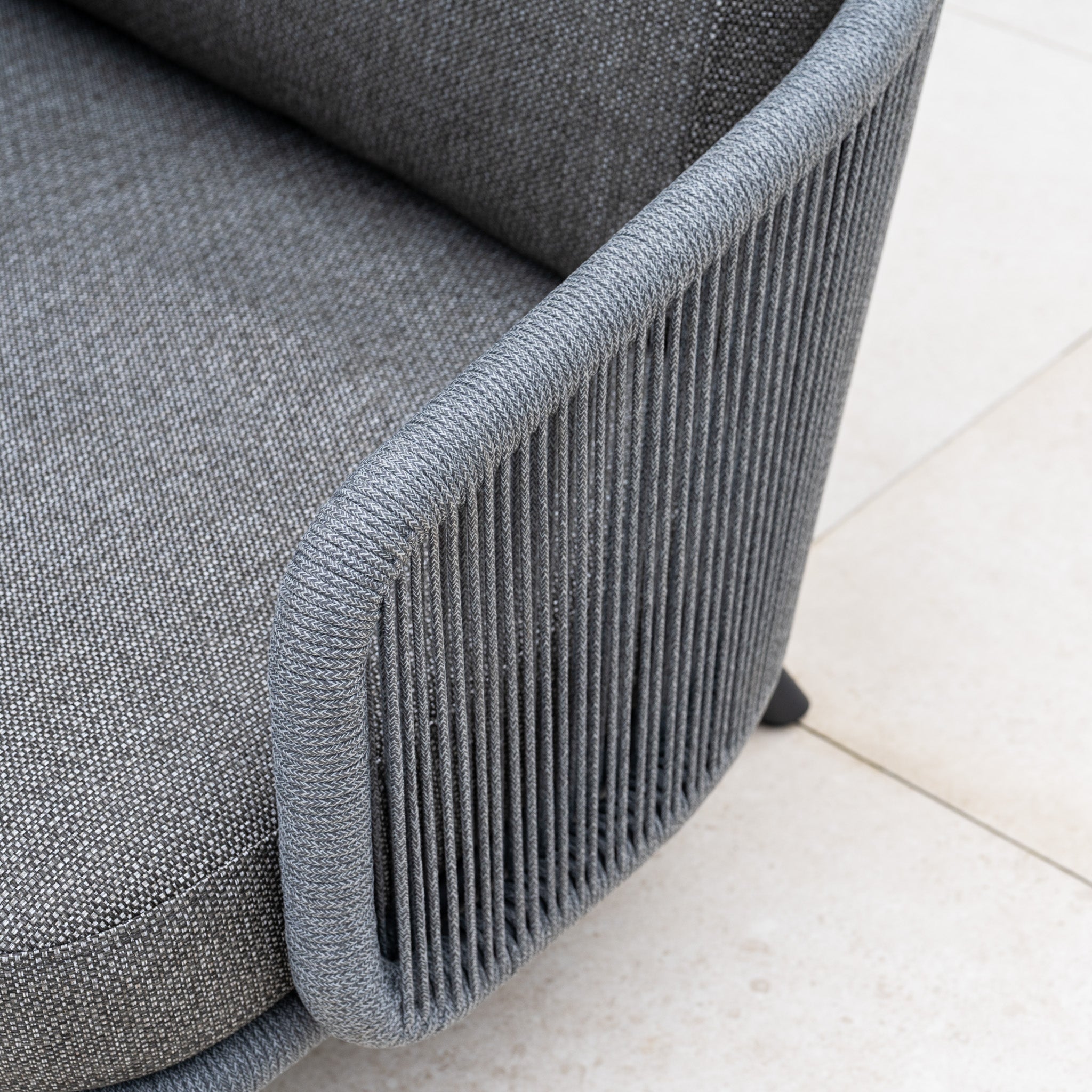 Close-up of a modern gray upholstered chair with a woven rope armrest, stylishly paired with the Dekota Large Corner Sofa Dining Set with Altura Rising Table in Charcoal, set against a light tile floor background.