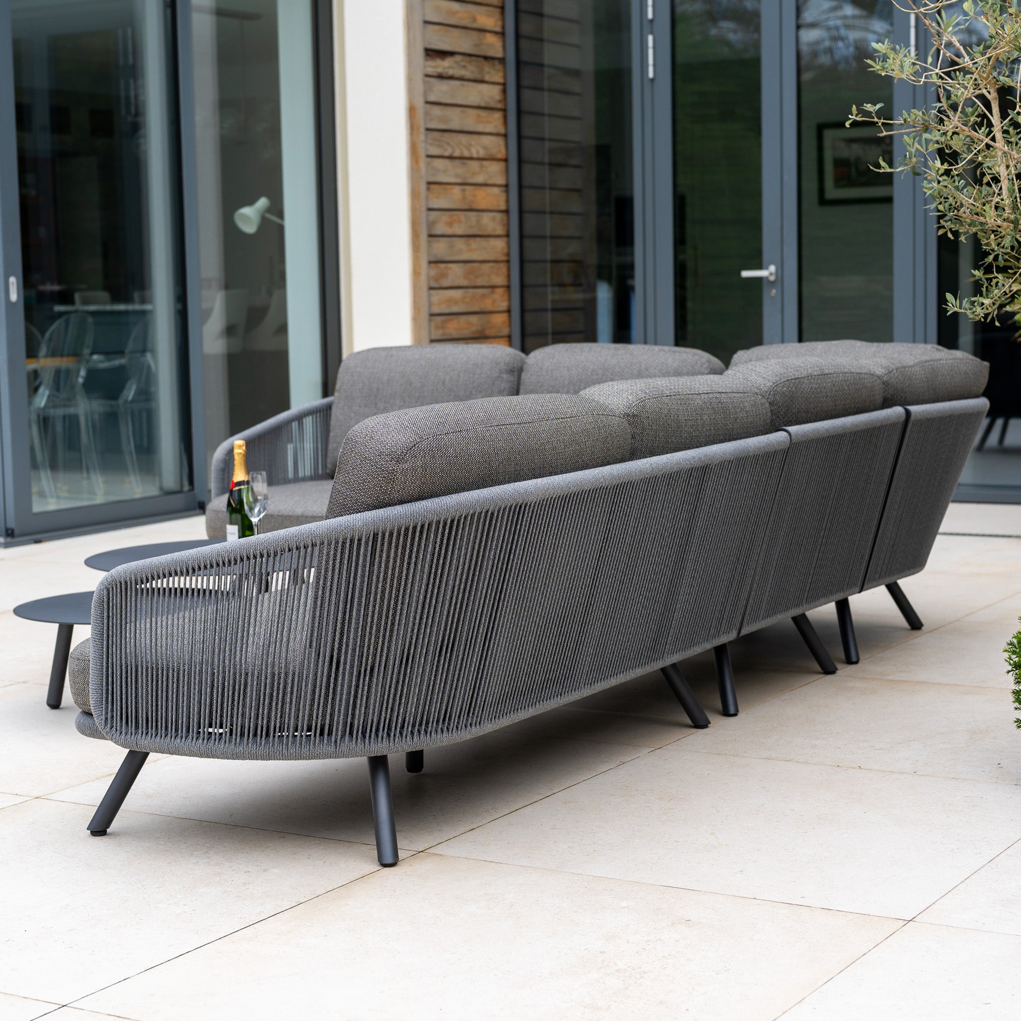 A Dekota Large Corner Sofa Set in Charcoal sits elegantly on the patio as outdoor furniture, accompanied by a bottle of champagne and two glasses.