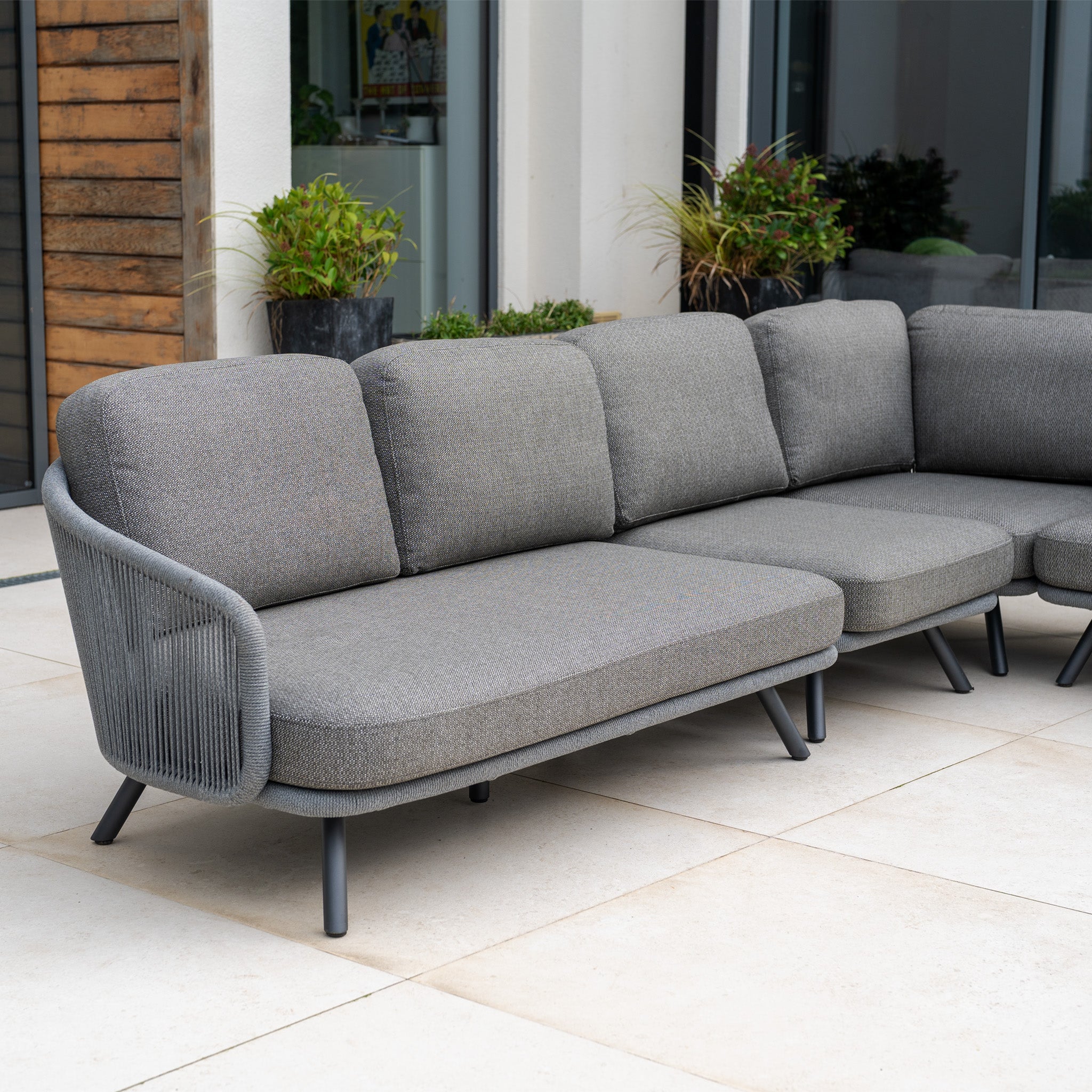 The Dekota Large Corner Sofa Set in Charcoal is a stylish outdoor sectional with cushions, perfect for patios. Its elegant look complements potted plants and large windows, enhancing any outdoor furniture arrangement.