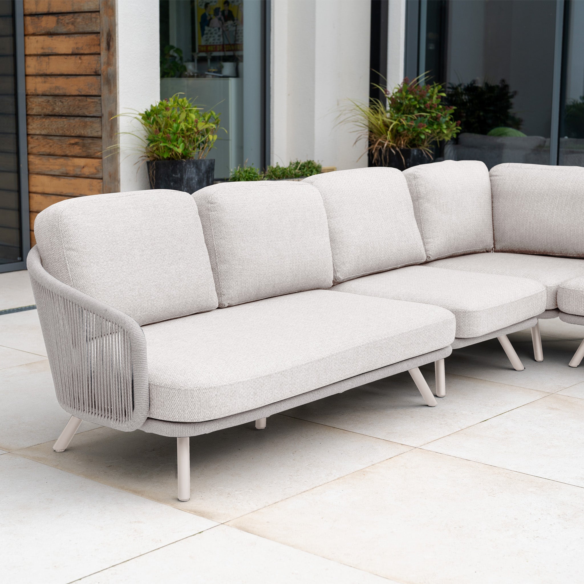 The Dekota Large Corner Sofa Set in Fawn features plush cushions and a hydrophobic rope weave, offering stylish outdoor seating for your tiled patio near potted plants and a modern house.