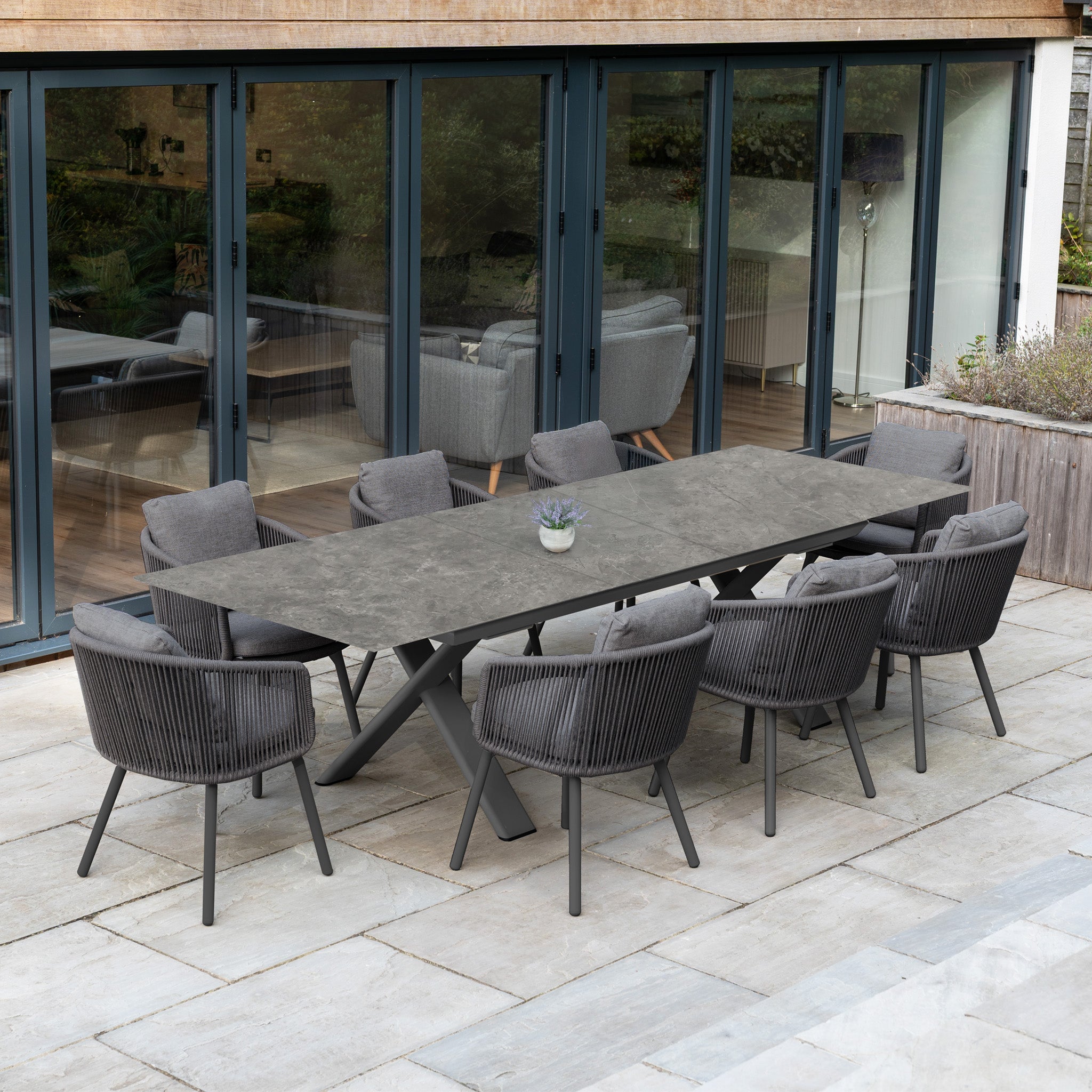 The Dekota 8 Seat Rope Dining Set with Albatross Extending Table in Charcoal sits on a stone patio beside glass doors, featuring stylish rope chairs for modern outdoor dining.