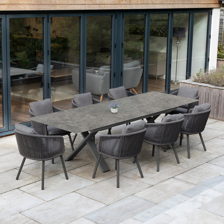 The Dekota 8 Seat Rope Dining Set with Albatross Extending Table in Charcoal sits on a stone patio beside glass doors, featuring stylish rope chairs for modern outdoor dining.
