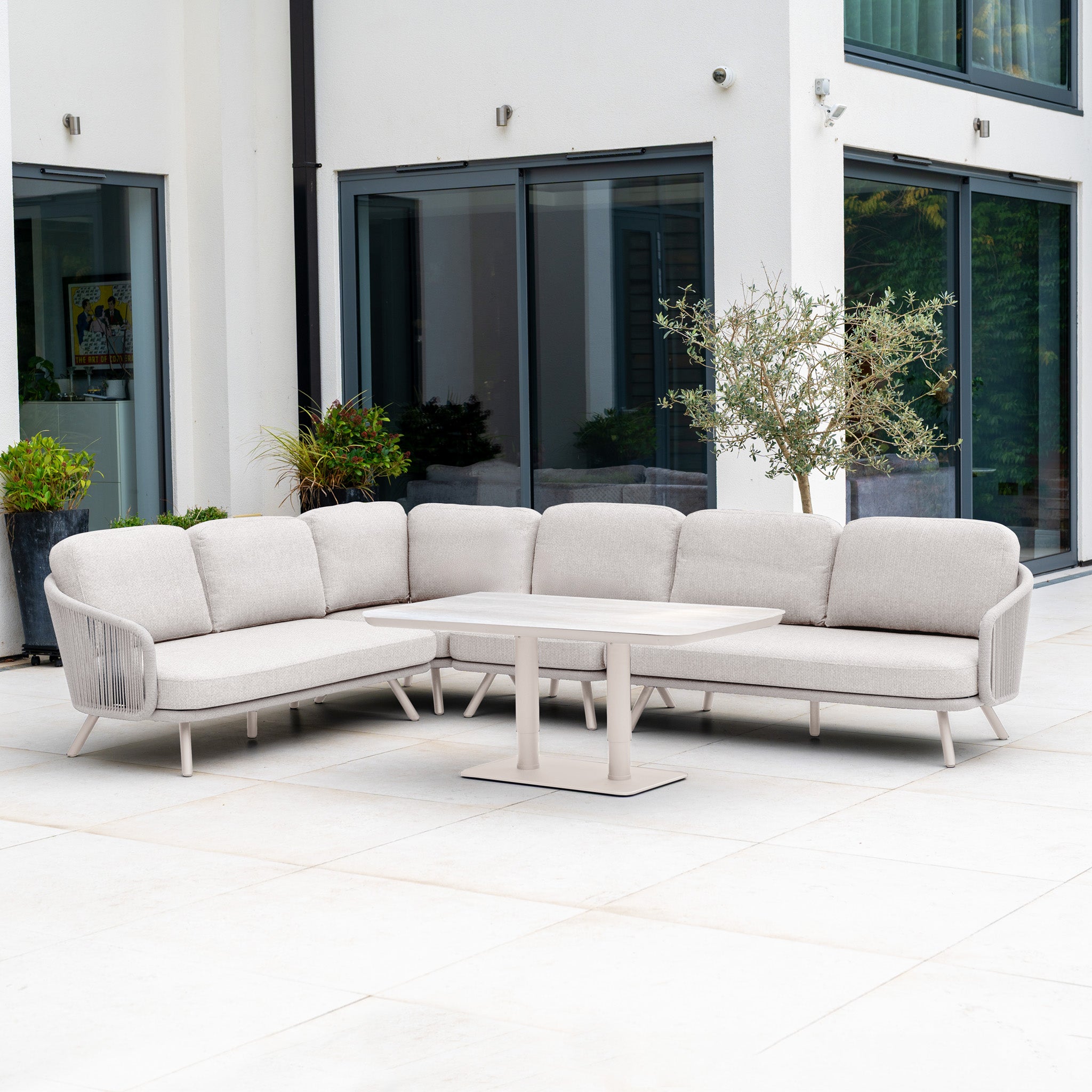 The Dekota Large Corner Sofa Dining Set with Altura Rising Table in Fawn sits on a spacious modern patio, surrounded by plants and large windows.