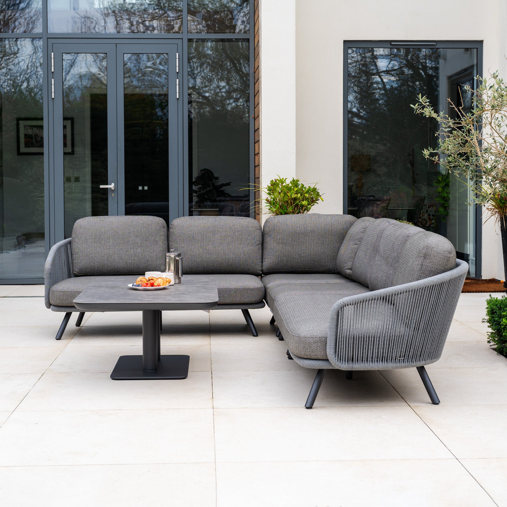 Modern outdoor patio featuring the Dekota Large Corner Sofa Dining Set with Altura Rising Table in Charcoal, set on a tiled terrace with snacks.