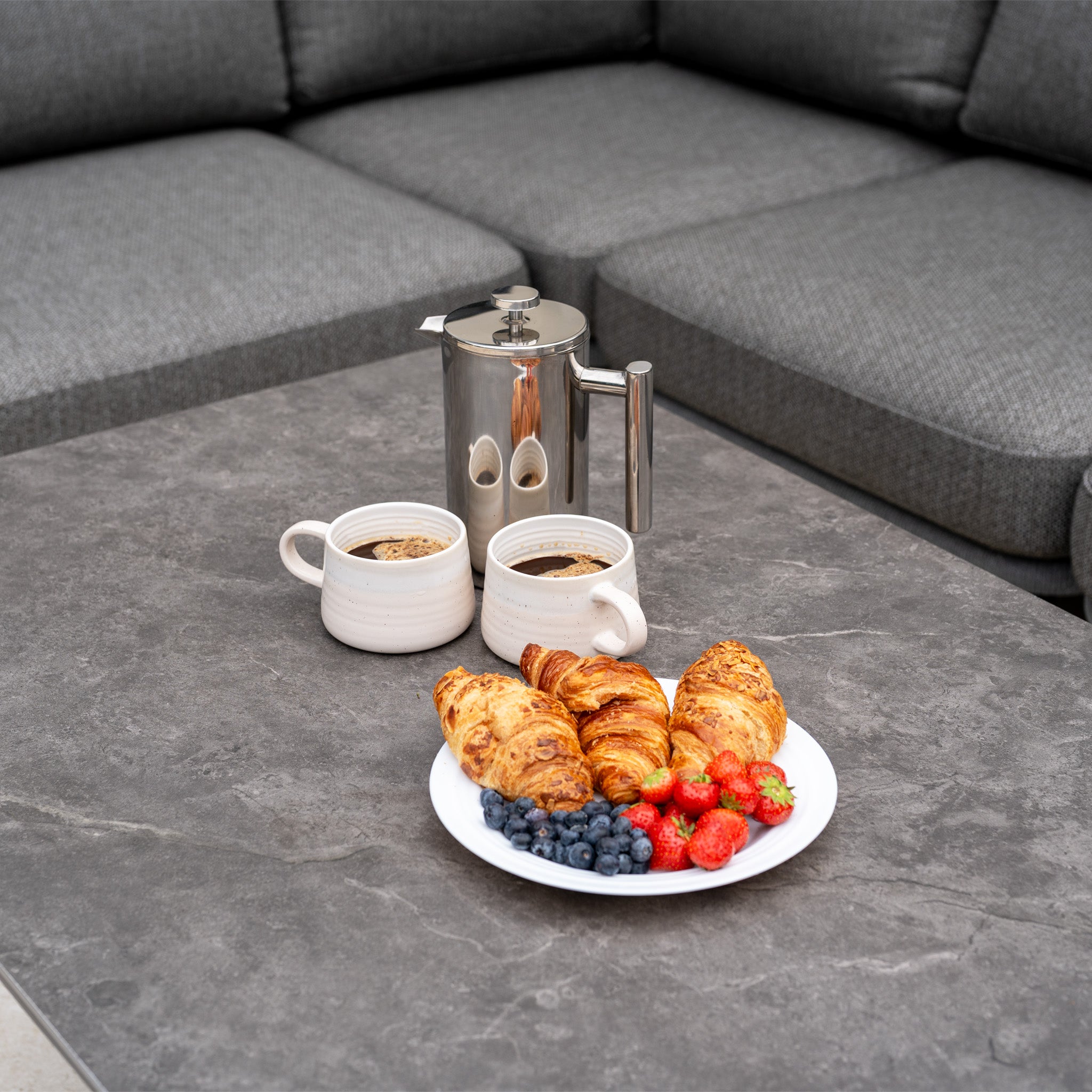 A plate with croissants, strawberries, blueberries, two mugs of coffee, and a coffee pot rests on the gray Altura Rising Table beside the Dekota Large Corner Sofa Dining Set in Charcoal.