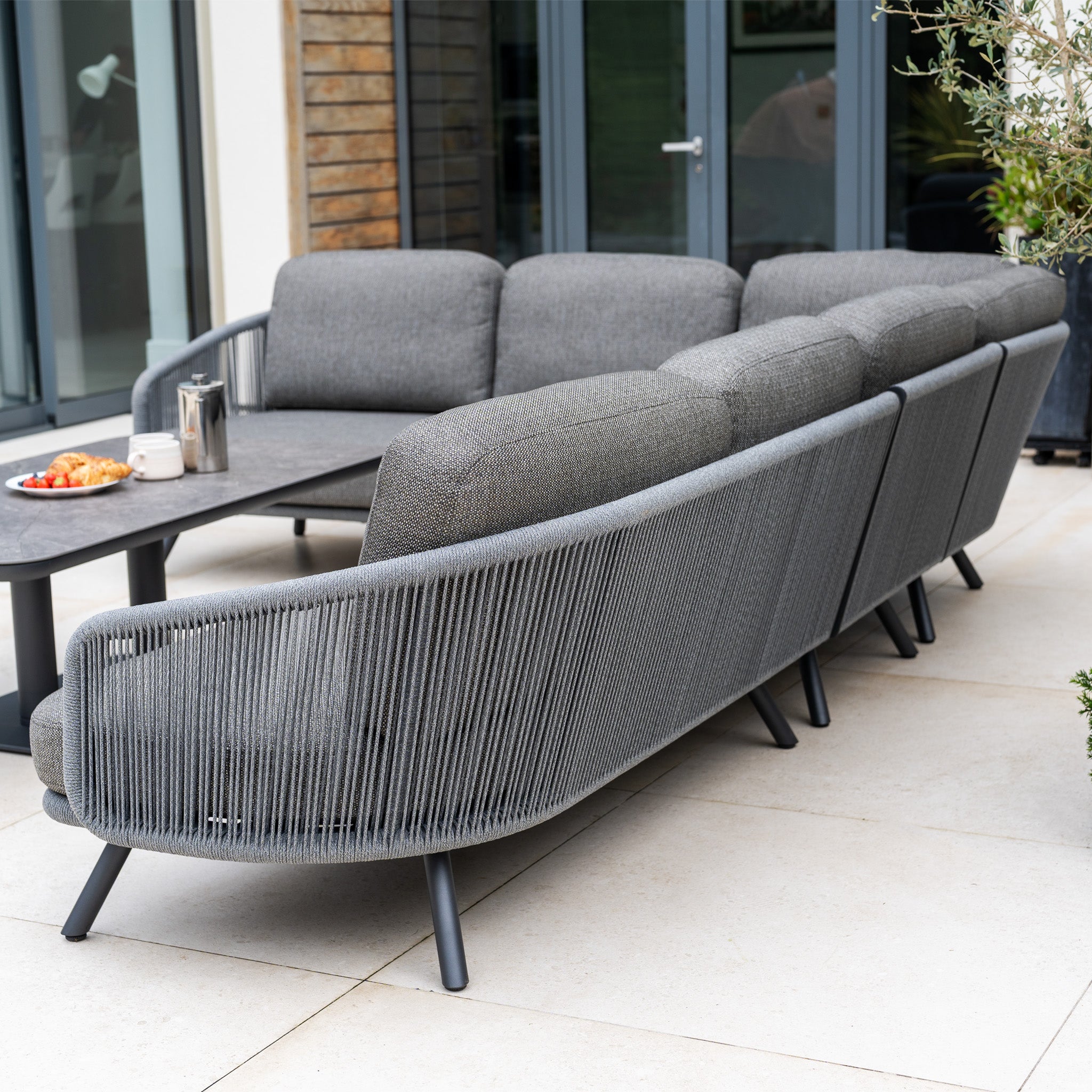 The Dekota Large Corner Sofa Dining Set with Altura Rising Table in Charcoal sits on a patio near glass doors, complete with snacks and a teapot on the table.