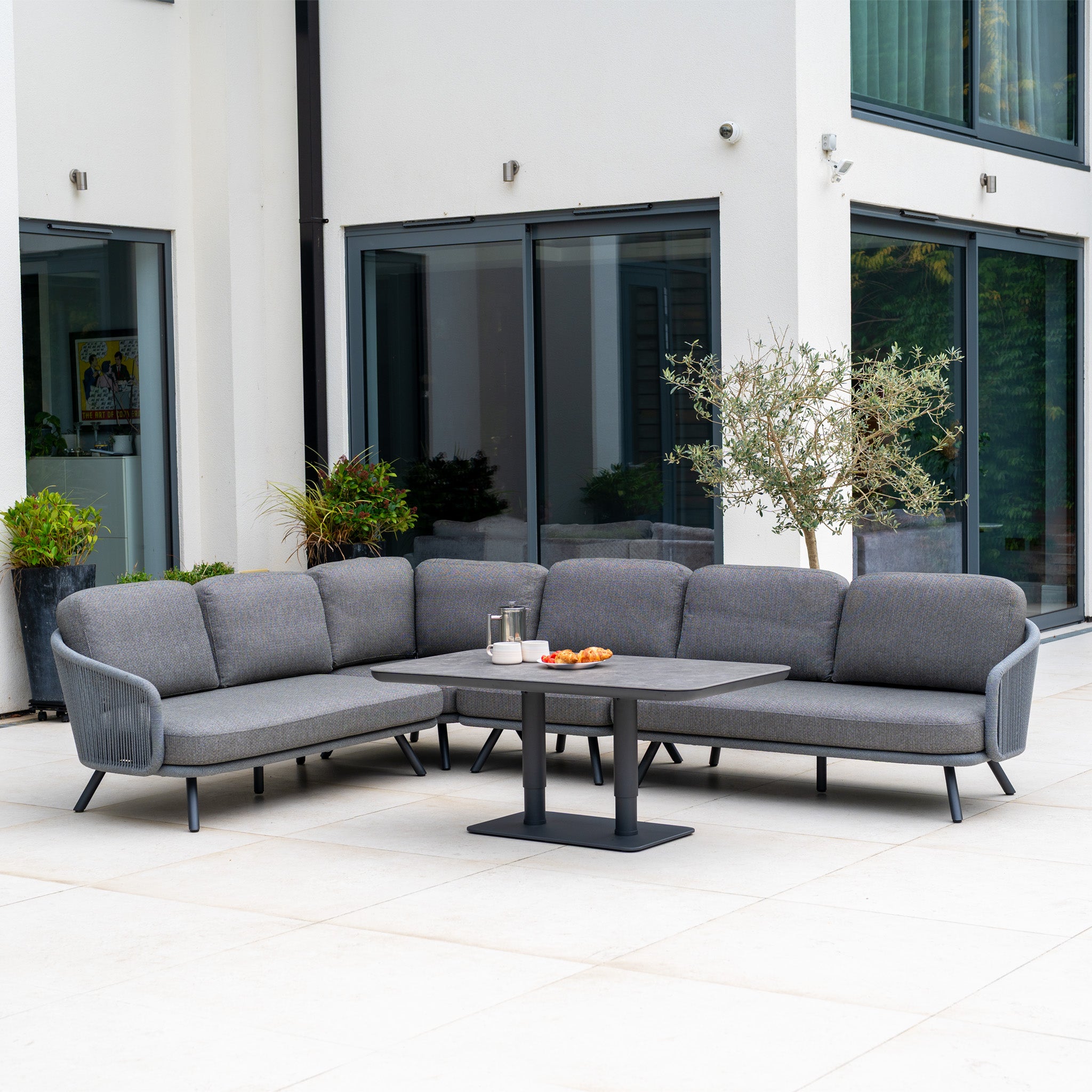 Modern patio featuring the Dekota Large Corner Sofa Dining Set with Altura Rising Table in Charcoal, surrounded by lush plants and snacks, outside a white building with expansive windows.