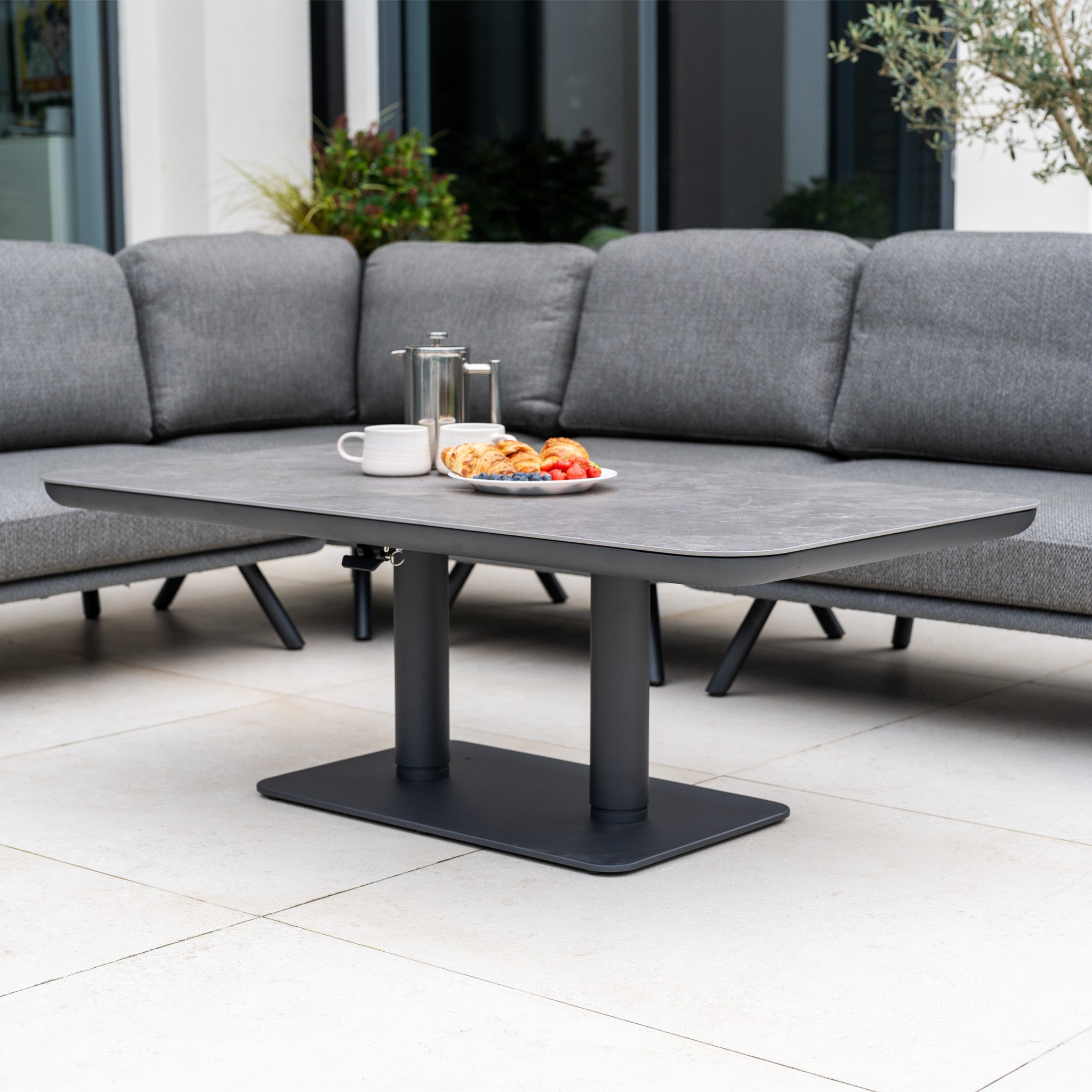 The Dekota Large Corner Sofa Dining Set with Altura Rising Table in Charcoal features a modern grey rising dining table with pastries, berries, and coffee, surrounded by a spacious charcoal outdoor sofa.
