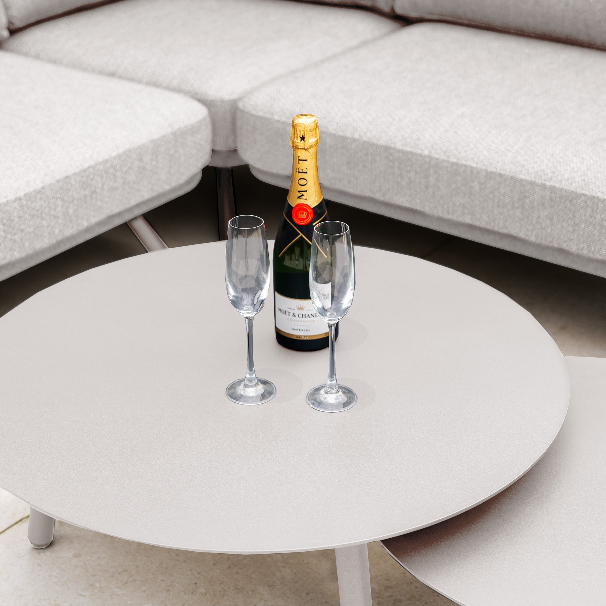 A bottle of Moët & Chandon champagne and two empty glasses rest on a round table in front of the Dekota Corner Sofa Set in Fawn, a chic gray outdoor sofa crafted with hydrophobic rope weave for stylish lounging.
