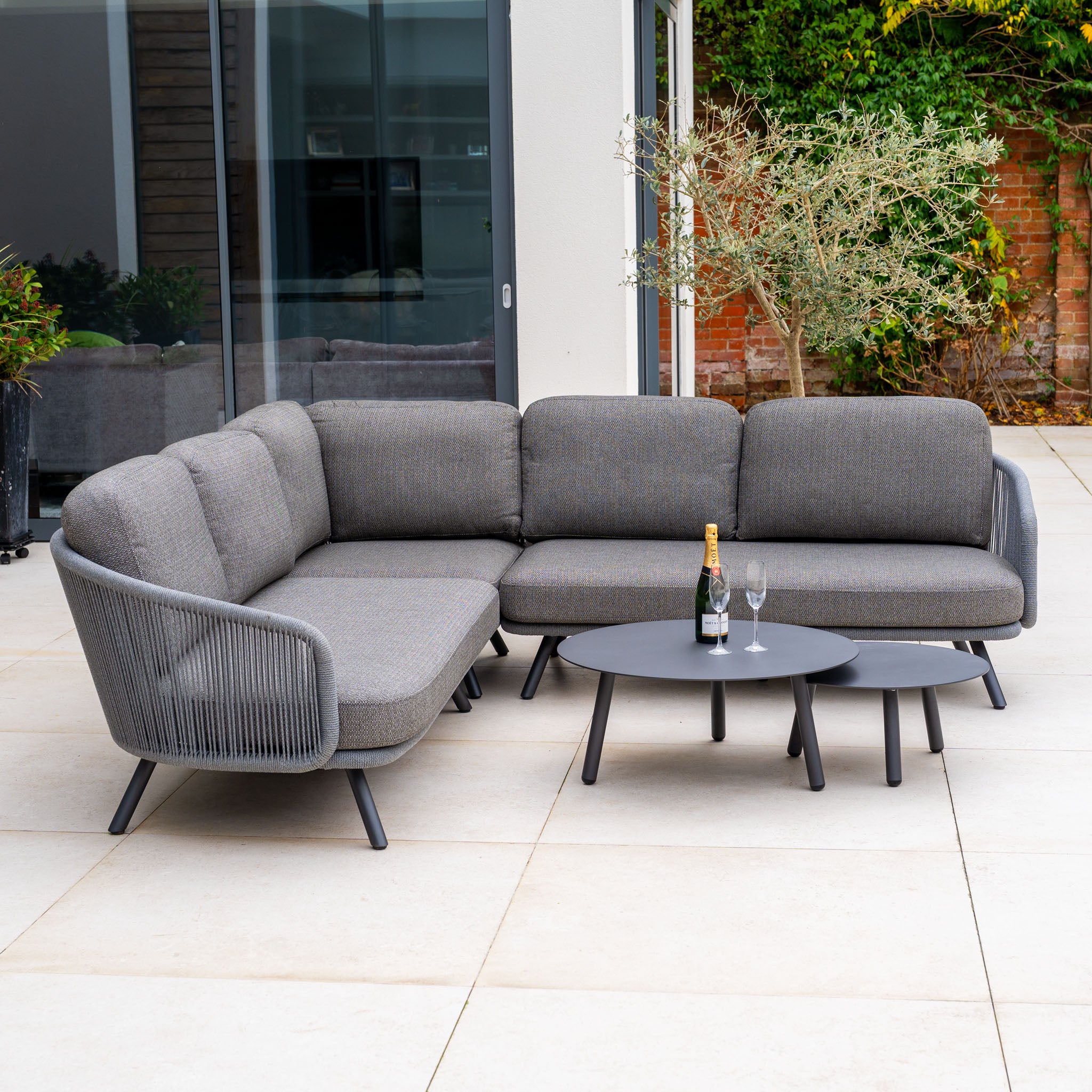 Dekota Corner Sofa Set in Charcoal, featuring hydrophobic rope weave, two round black tables, champagne bottle, and glasses on a tiled patio.
