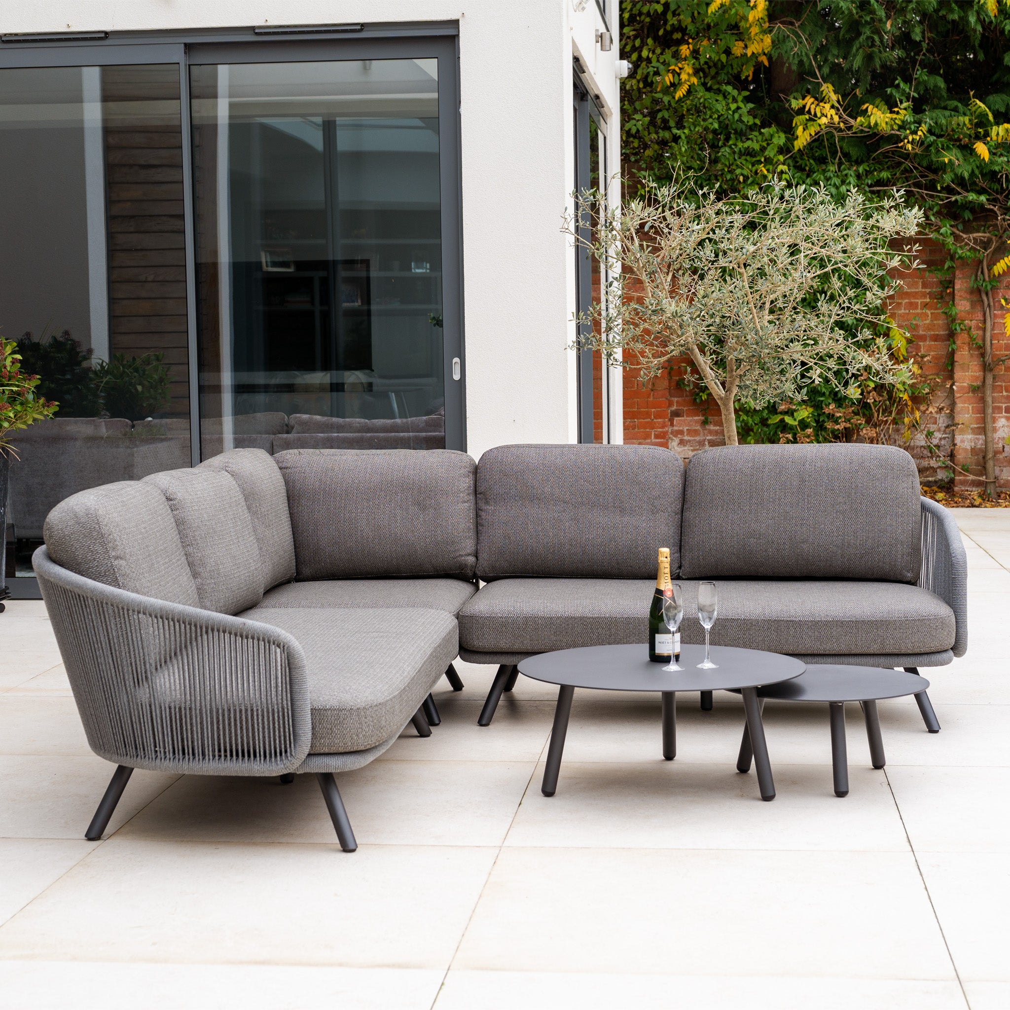 The Dekota Corner Sofa Set in Charcoal, with champagne and glasses on tables, features a hydrophobic rope weave and is displayed on a patio beside a modern building.