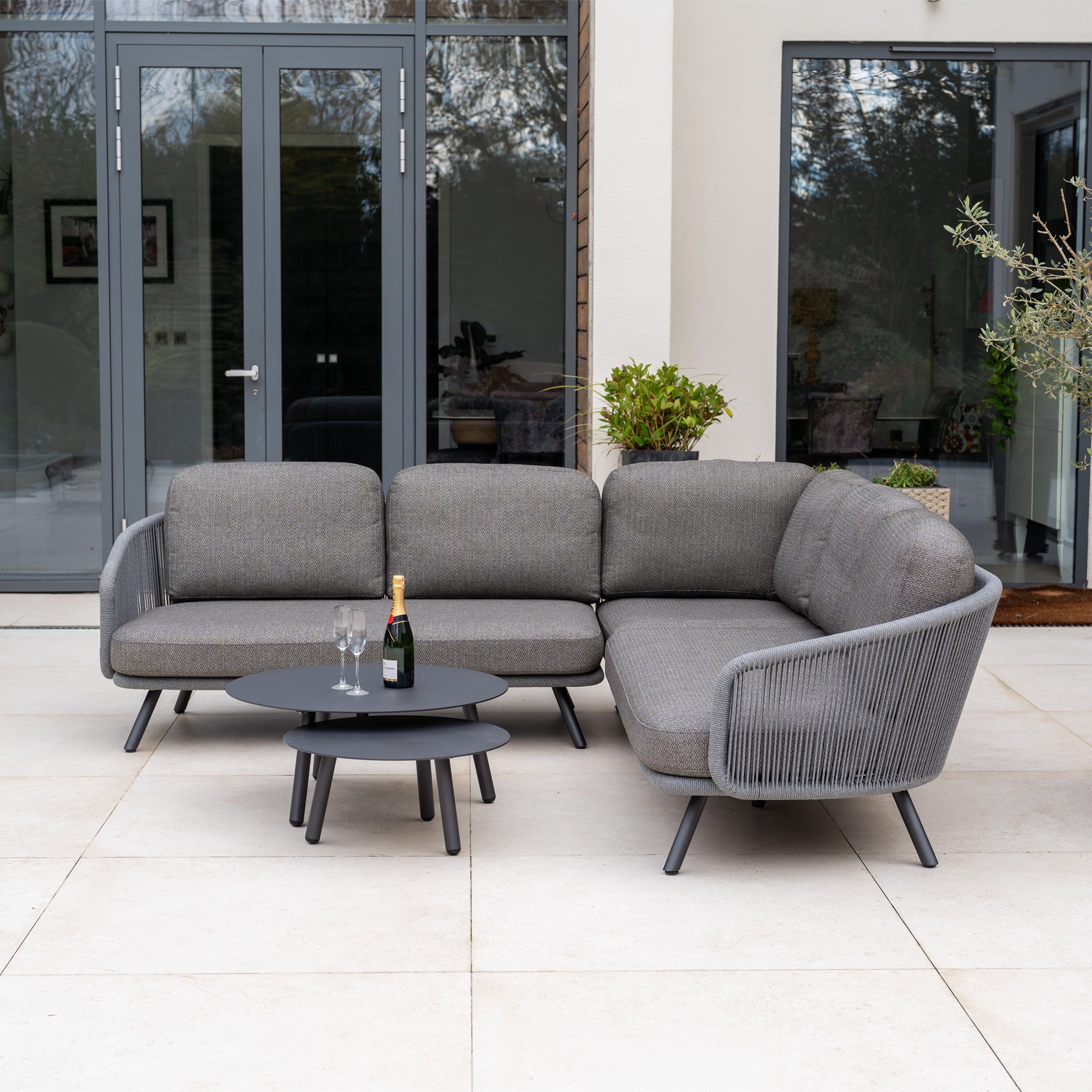 Dekota Corner Sofa Set in Charcoal with a round coffee table, champagne, and glasses on a patio by glass doors, featuring hydrophobic rope weave for all-weather comfort.