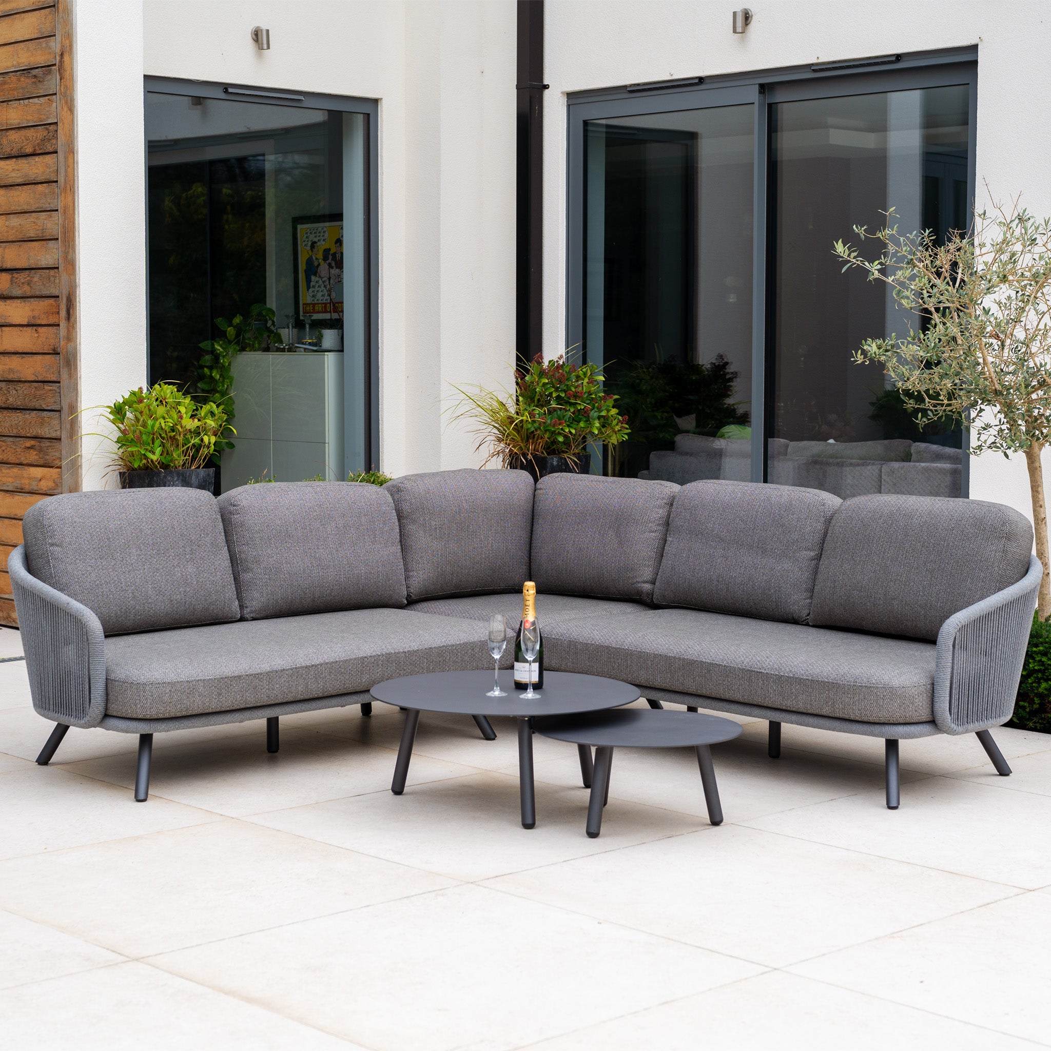 The Dekota Corner Sofa Set in Charcoal features hydrophobic rope weave details and comes with two round tables, creating a modern, stylish patio setup perfect for relaxing with champagne and glasses.