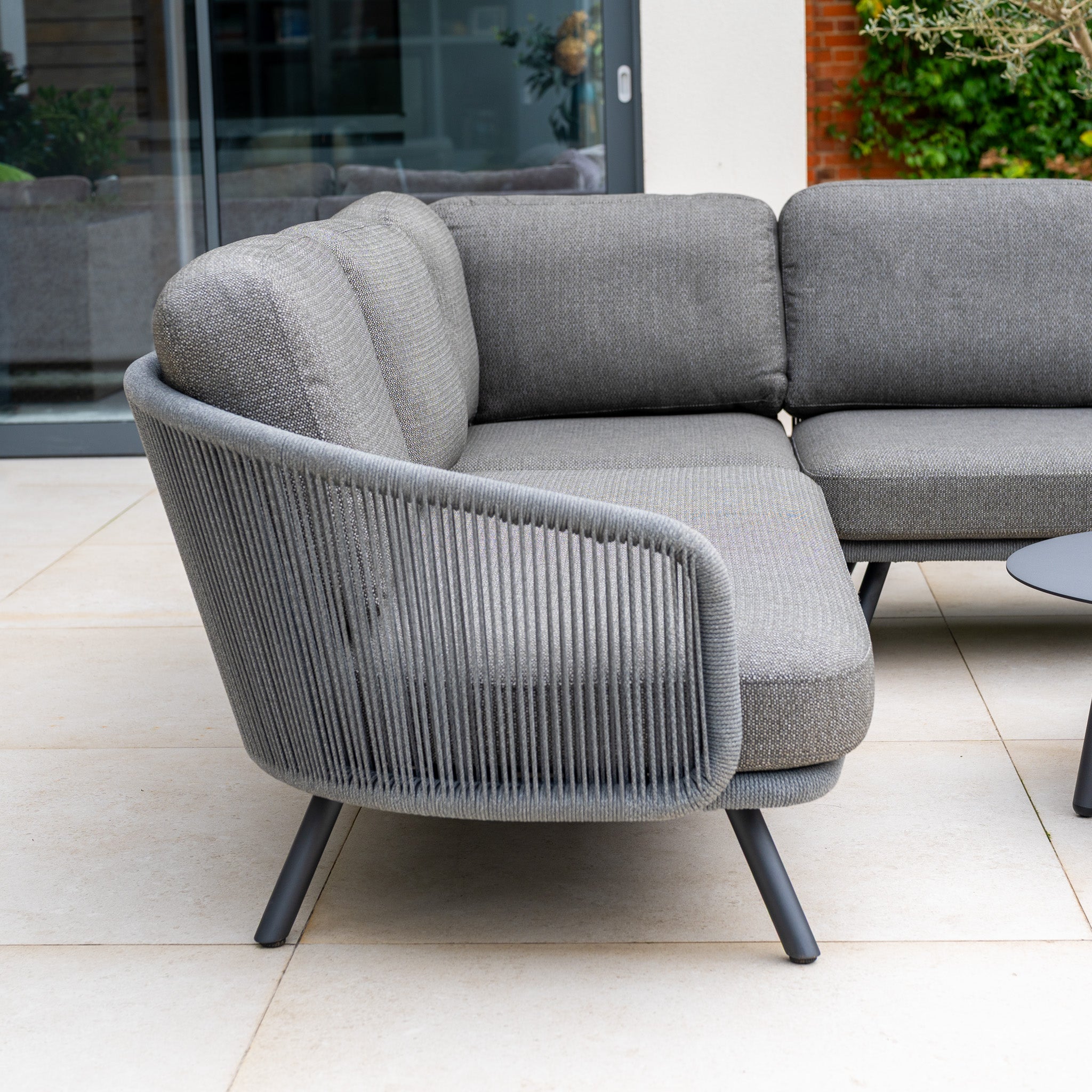 The Dekota Corner Sofa Set in Charcoal with hydrophobic rope weave sits on a tiled patio beside a round black table.