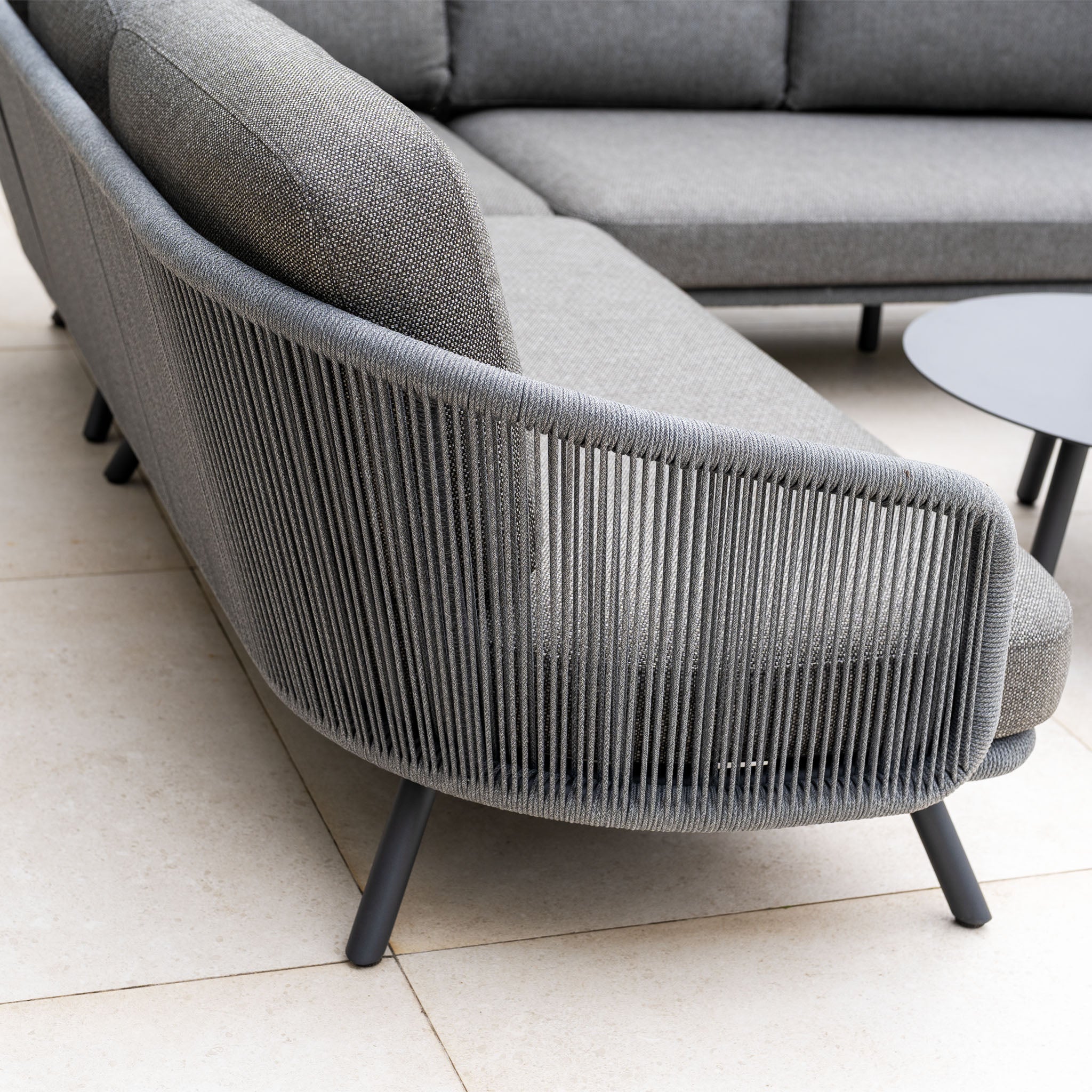 Dekota Corner Sofa Set in Charcoal features hydrophobic rope weave detailing on the back and comes with a round black side table, shown on tiled flooring.