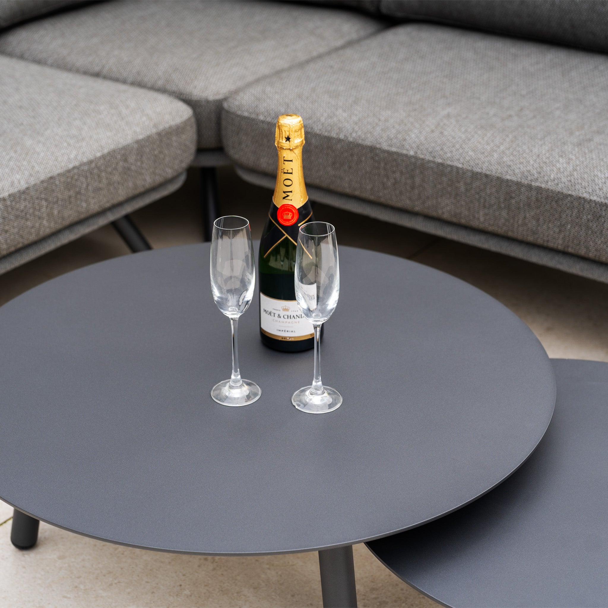 A bottle of champagne and two empty flutes on a round gray table in front of the Dekota Corner Sofa Set in Charcoal, featuring elegant hydrophobic rope weave details.