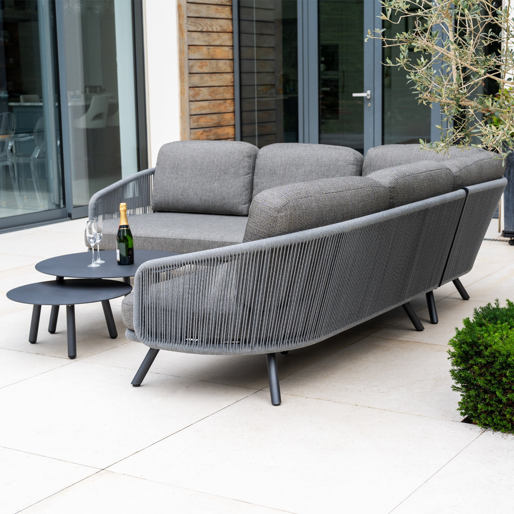Modern outdoor patio featuring a Dekota Corner Sofa Set in Charcoal with hydrophobic rope weave, two round tables, wine glasses, and champagne.