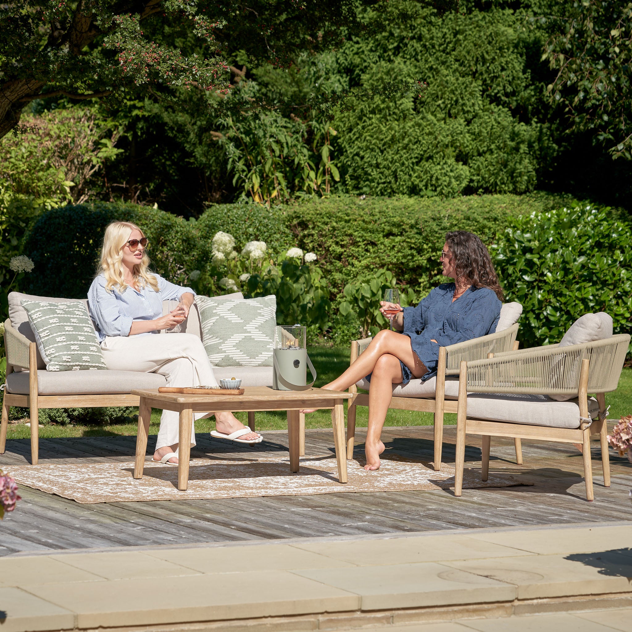 Two women sit chatting on the elegant Pacific Lifestyle - Denver 2 Seat Sofa Set in Sage Green in a lush garden, surrounded by vibrant greenery and flowers.