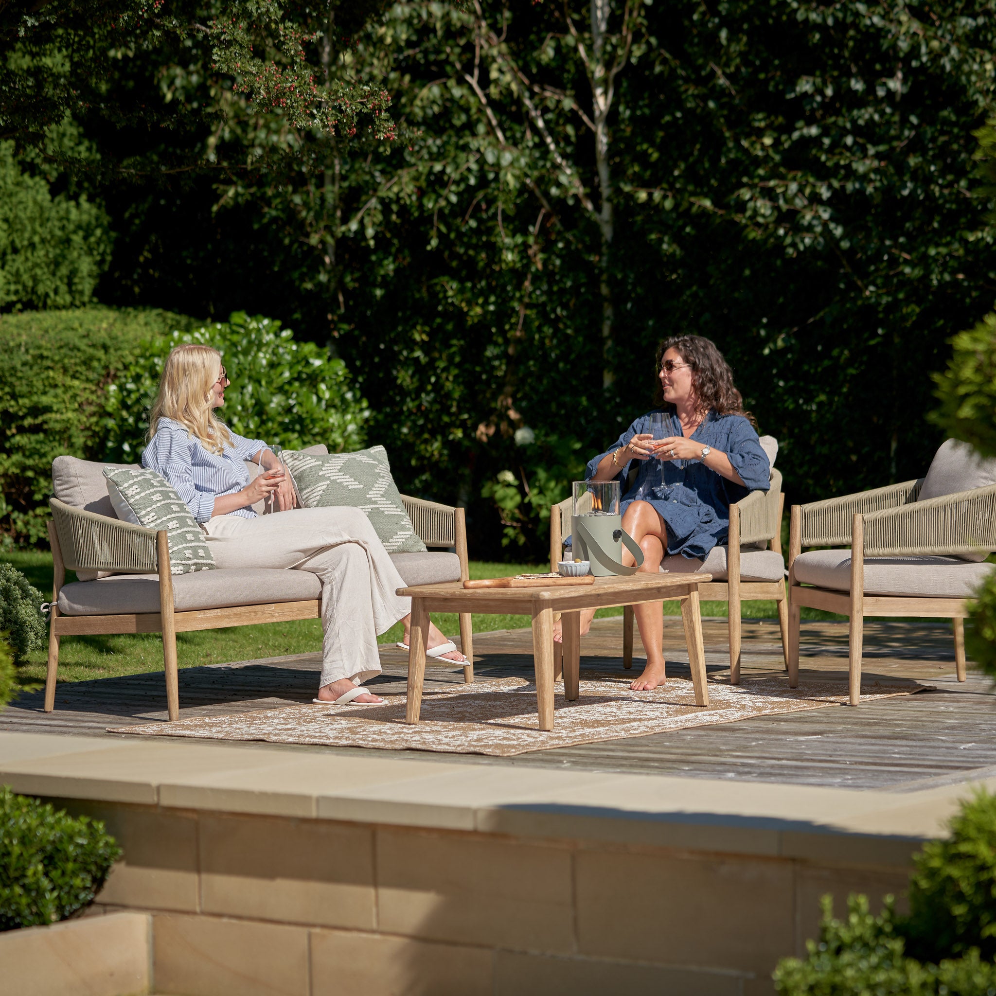 Two people relax on the Pacific Lifestyle - Denver 2 Seat Sofa Set in Sage Green, chatting amid lush greenery. The elegant acacia wood furniture enhances the serene atmosphere on their sunny patio.