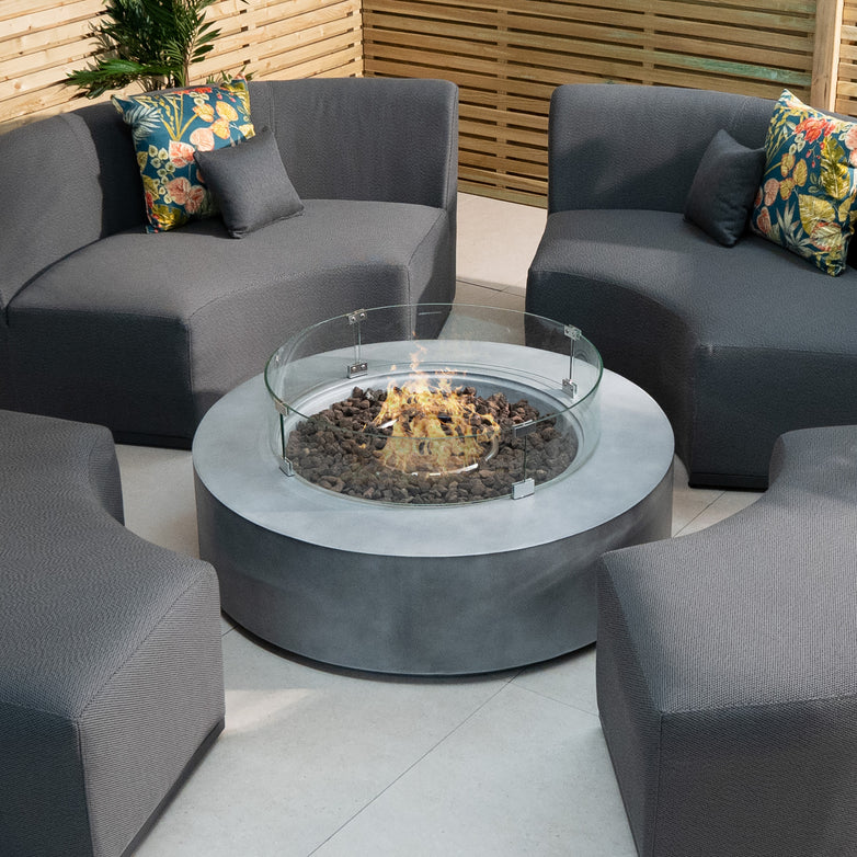 Modern outdoor seating area with gray cushioned chairs around an Ember Round Firepit Coffee Table in Stone Grey, decorated with vibrant pillows. This fire pit, made from high-quality 304 stainless steel, provides a cozy ambiance with its impressive 14kW heat output.