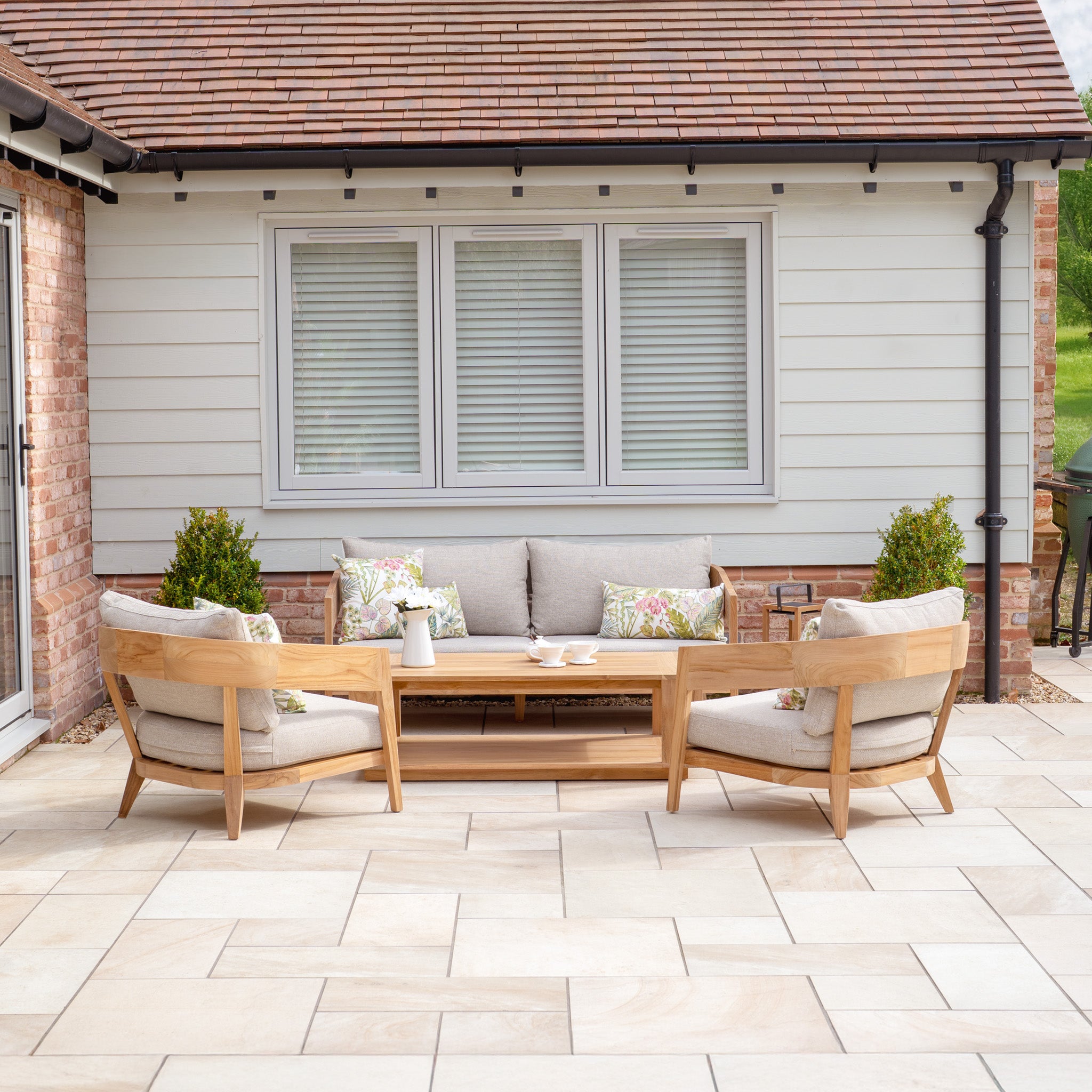 The patio boasts wooden furniture with plush cushions and potted plants for inviting garden décor, complemented by the Enna 3 Seat Sofa Set with Coffee Table in Fawn, enhancing comfort against a house with shuttered windows.