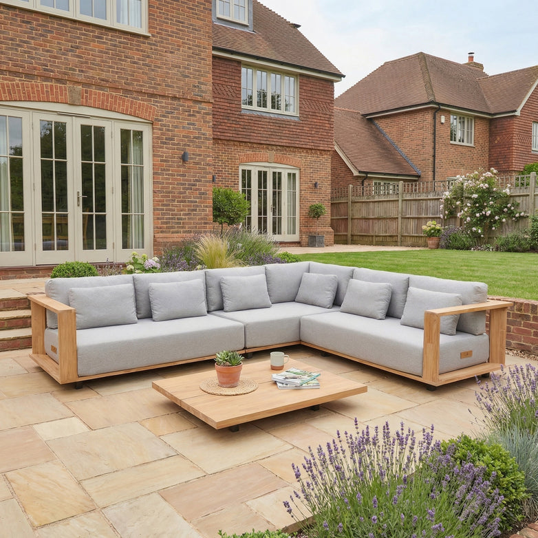 Modern outdoor patio with the Eternity Right Hand Teak Corner Group in Light Grey, an L-shaped sofa and low table, set in a landscaped garden beside a brick house.