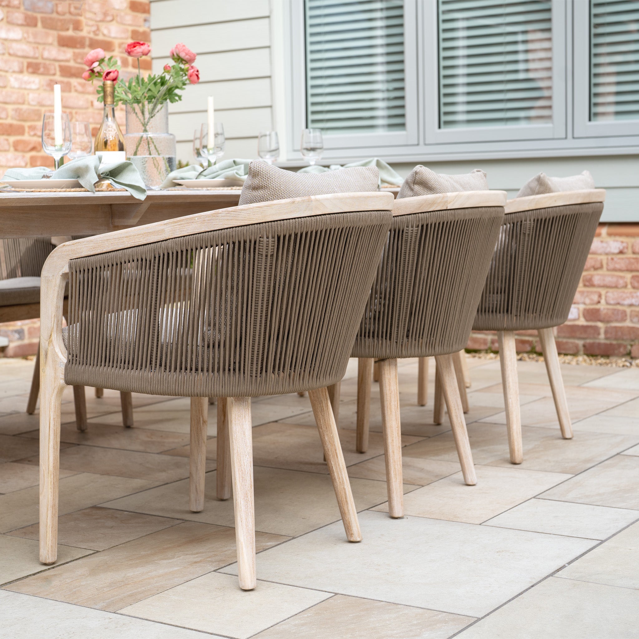 An outdoor patio showcases the Evora 8 Seat Rectangular Dining Set in Mocha, featuring a teak dining table and wicker chairs with mocha rope detailing. A brick wall and windows enhance the charm, highlighting the elegant place settings.