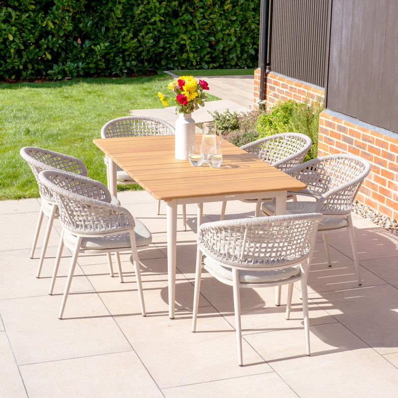 The Kalama 6 Seat Rectangular Dining Set in Latte showcases a beautiful teak table paired with six chic wicker chairs. A vase of flowers and two glasses grace the tabletop, enhancing its welcoming outdoor charm. Made from weather-resistant aluminum, this dining set offers both durability and elegance for any environment.