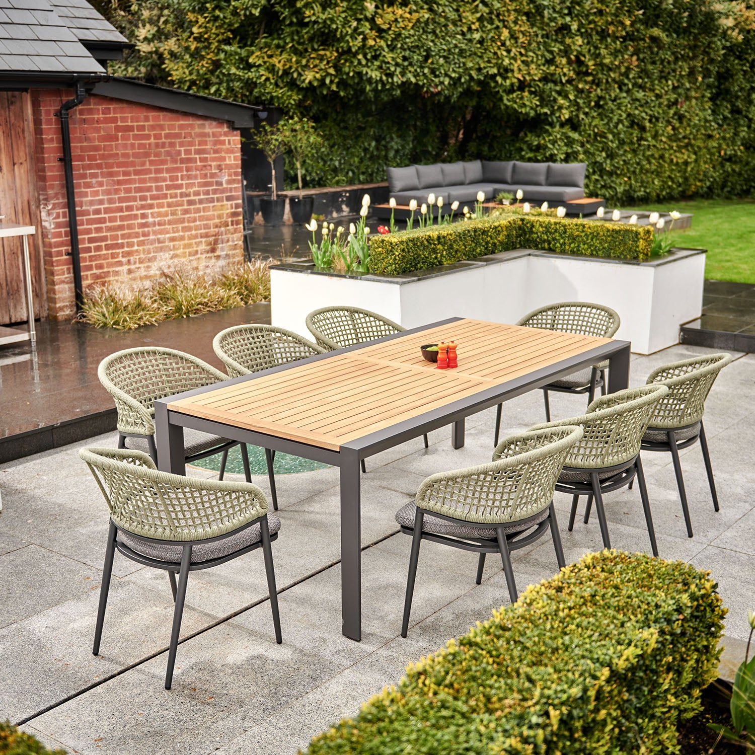 Outdoor patio featuring the Kalama 8 Seat Rectangular Extending Dining Set with a Teak Table in Olive Green, surrounded by greenery and set against a brick building backdrop.