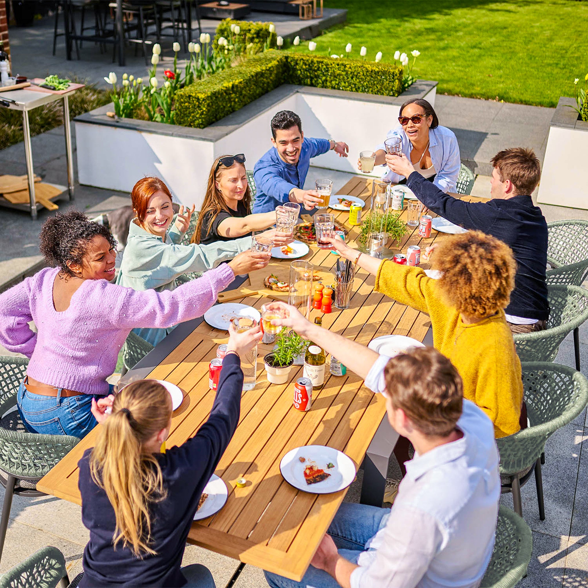 A diverse group of people sitting at an outdoor table, raising their glasses in a cheerful toast, enjoying their meal on a beautifully crafted Kalama 8 Seat Rectangular Extending Dining Set with a Teak Table in Olive Green, surrounded by bright weather and lush greenery.