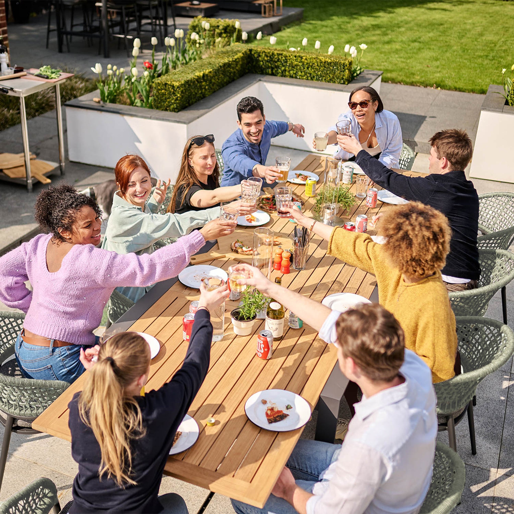 A group of friends clinked glasses around the Kalama 10 Seat Rectangular Extending Dining Set with a teak table in olive green, enjoying a meal together in a sunny backyard, seated comfortably on woven chairs.