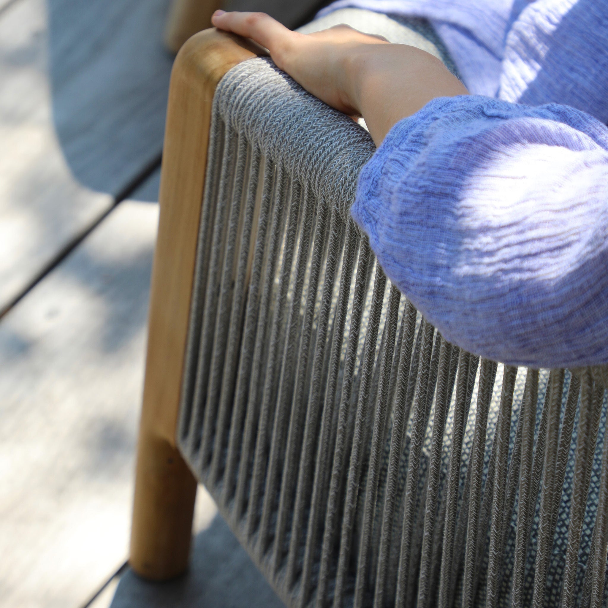 On a sunny day, a person’s hand rests on a wooden armrest of the Merano 2 Seat Sofa Set and Coffee Tables in Latte, featuring grey hydrophobic olefin rope weave detailing.