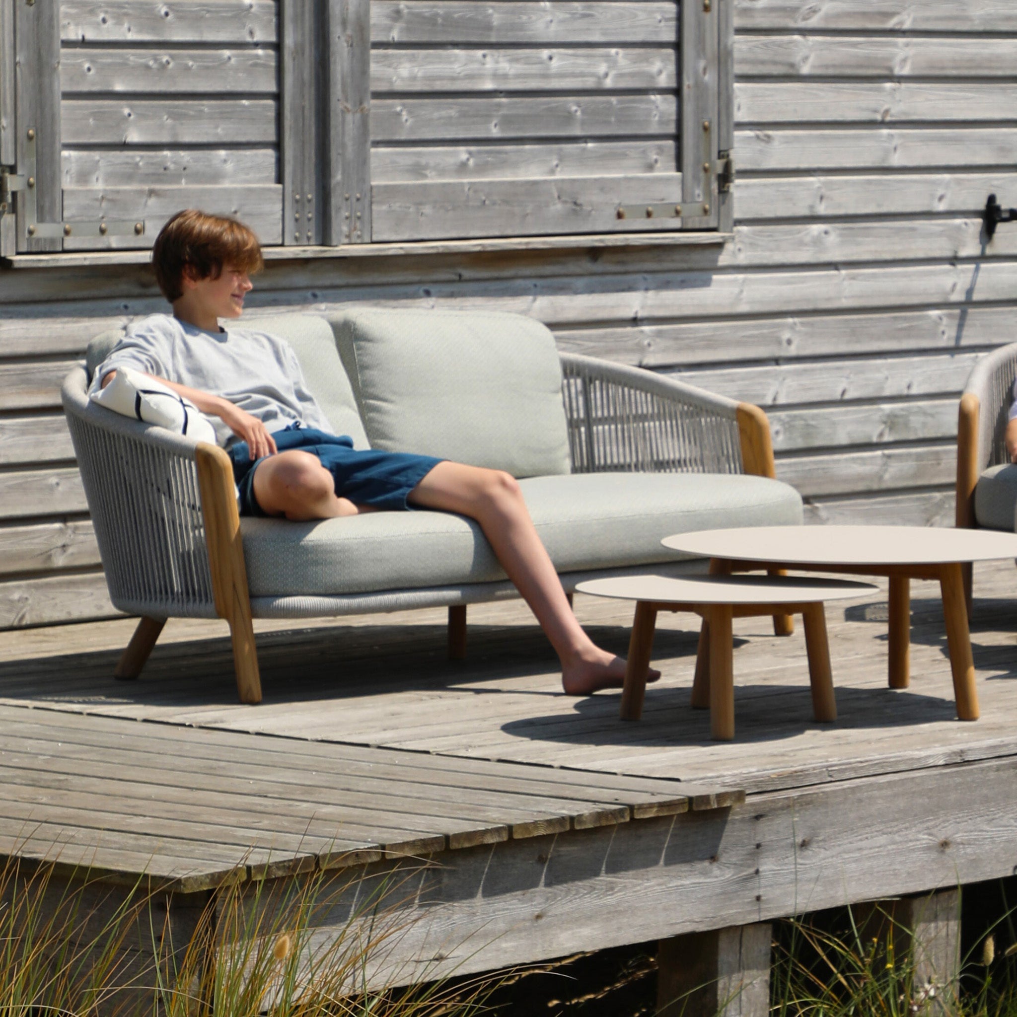 A person relaxes on a Merano 2 Seat Sofa Set and Coffee Tables in Latte, an epitome of outdoor luxury furniture, near a wooden building. This exquisite set features hydrophobic olefin rope weave accents and includes a round coffee table in front, with grassy plants below enhancing the serene ambiance.