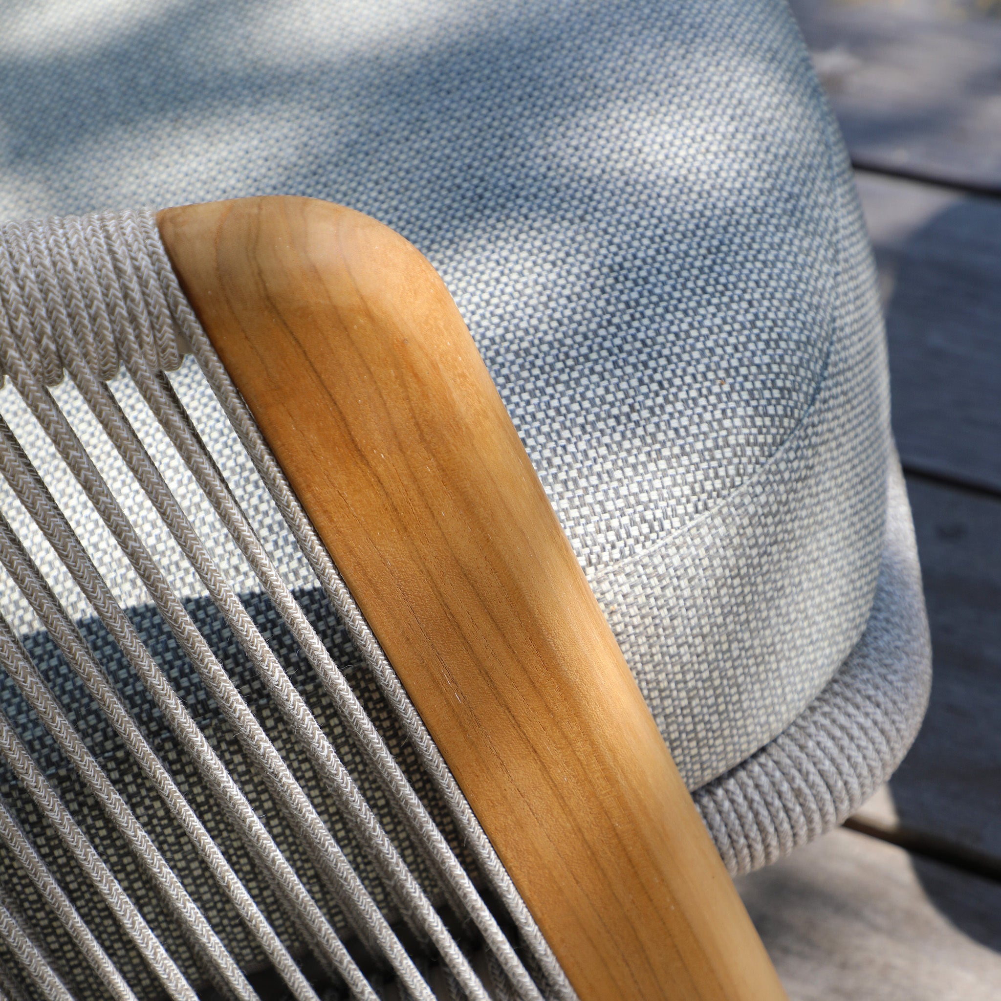 Close-up of a chair from the Merano 2 Seat Sofa Set and Coffee Tables in Latte, showcasing light gray fabric, wooden armrests, and hydrophobic olefin rope weave detail under the armrest, taken in sunlight.