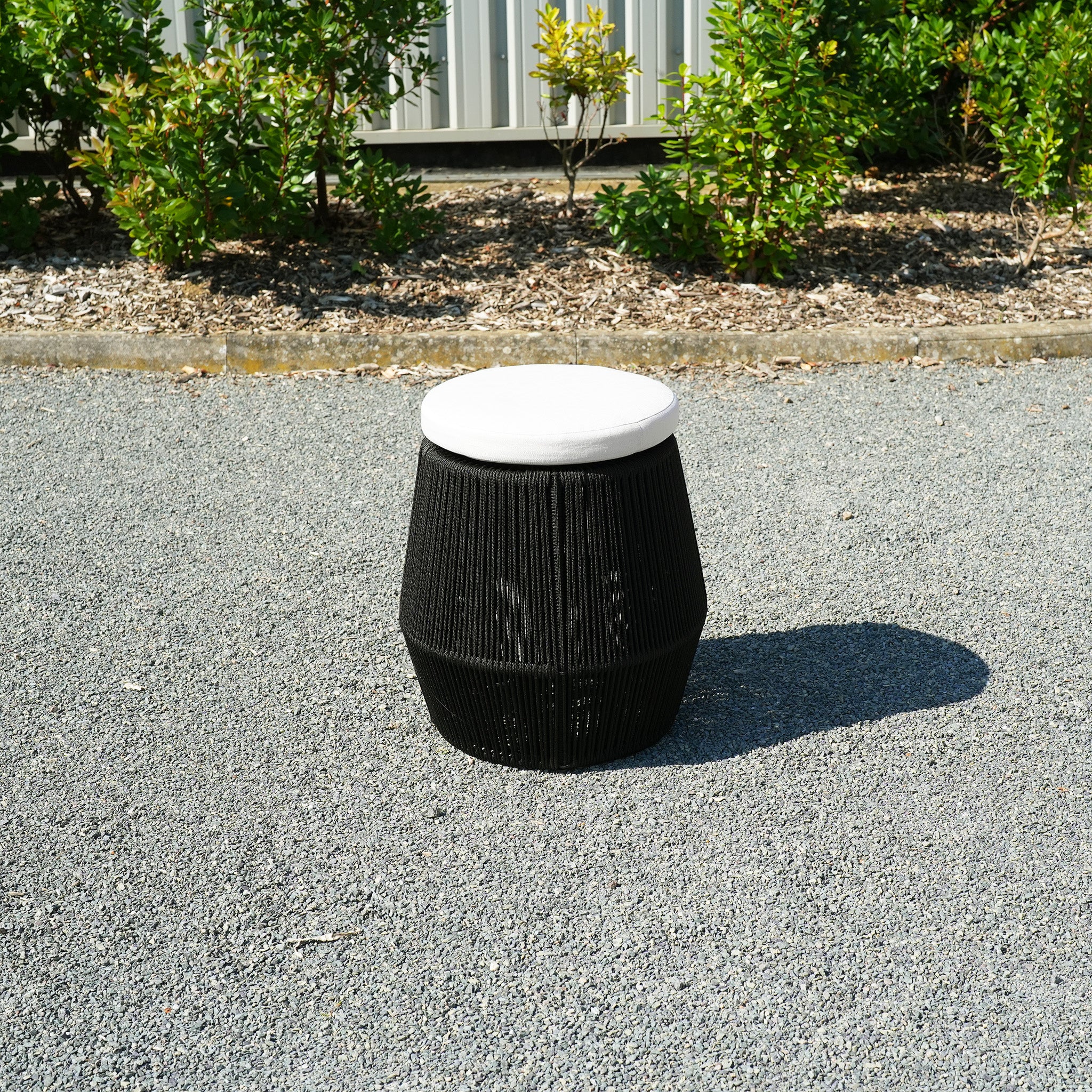 Rubin Stool in Black (Ex Display) with a white cushion on a gravel pathway, featuring an aluminium frame and black woven rope, surrounded by greenery and plants.