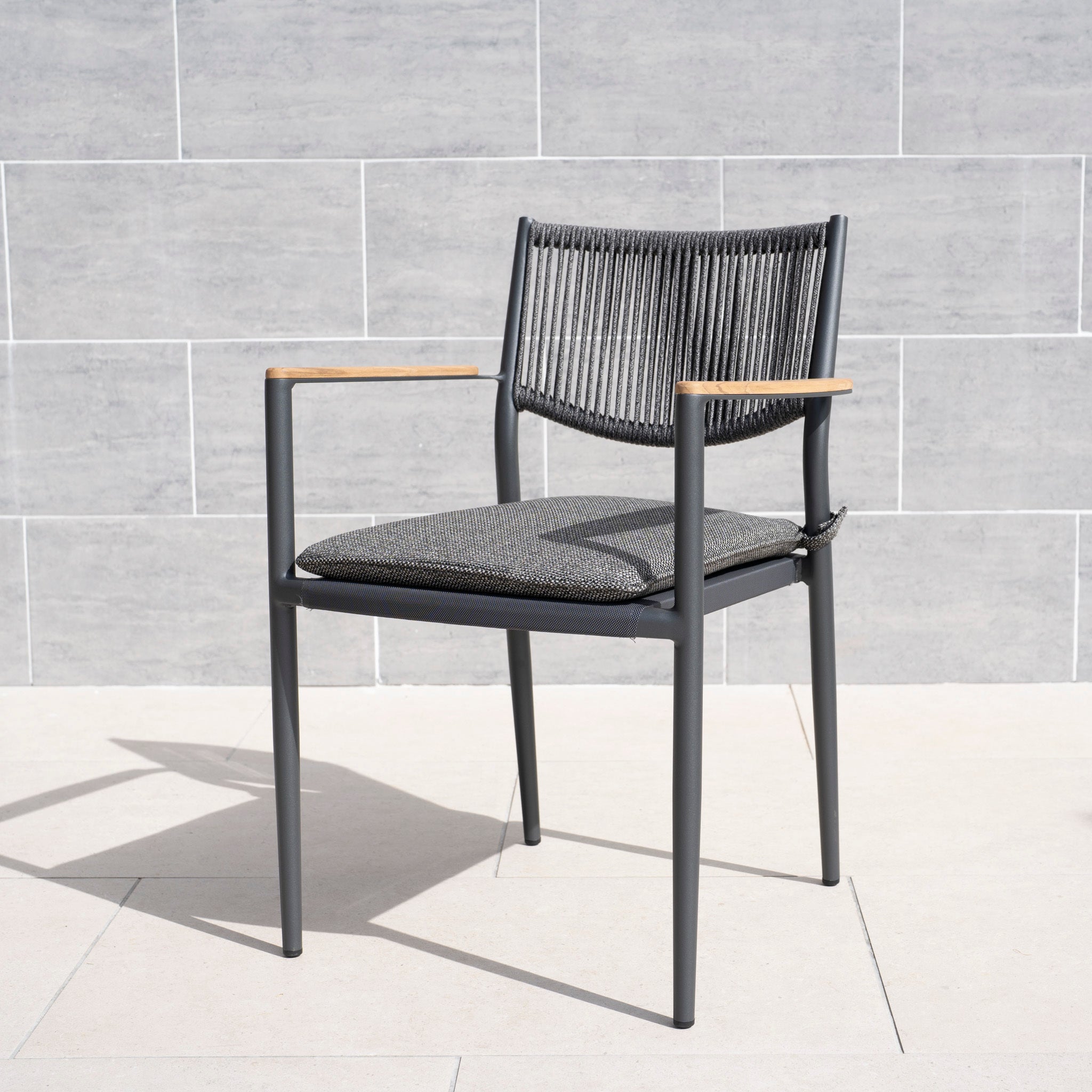 The Tellaro Rope Dining Chair in Grey (set of 6) features a woven back, weather-resistant cushion, and wooden armrests, shown here against a tiled wall.